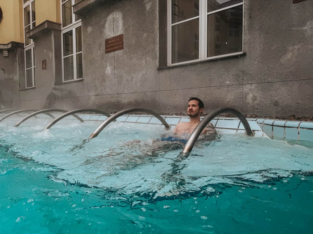 A man lies in the water under arched metal bars that release water jets in the thermal pool at Lukács Thermal Bath.