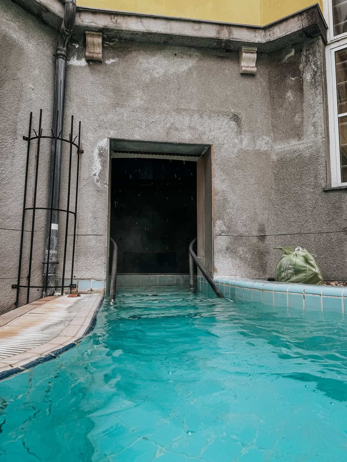 An outdoor pool area at Lukács Thermal Bath with a narrow passage leading into an indoor section, adjacent to a weathered gray wall.