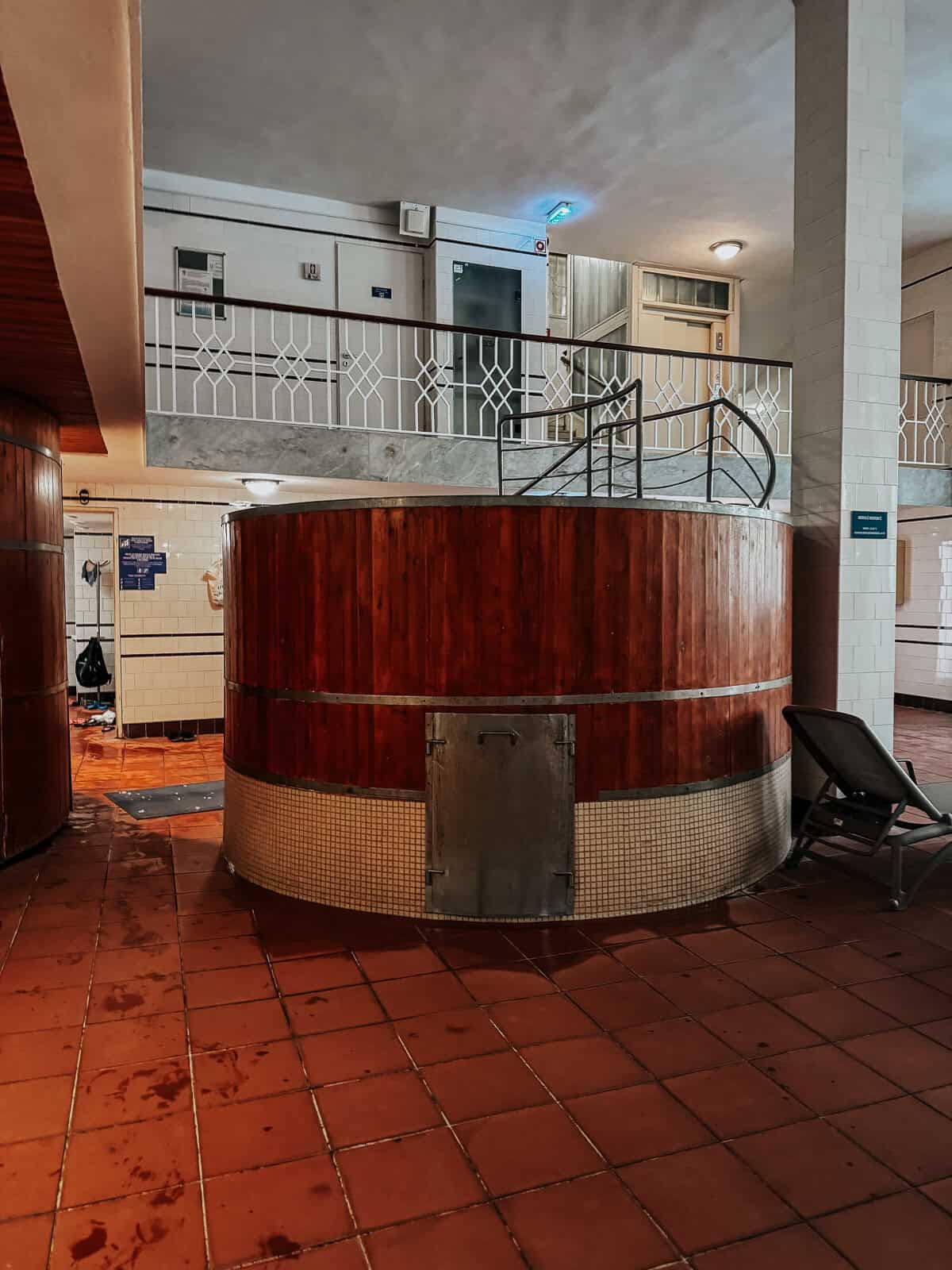 A large wooden hot tub in the interior of Lukács Thermal Bath, surrounded by tiled floors and metal railings.