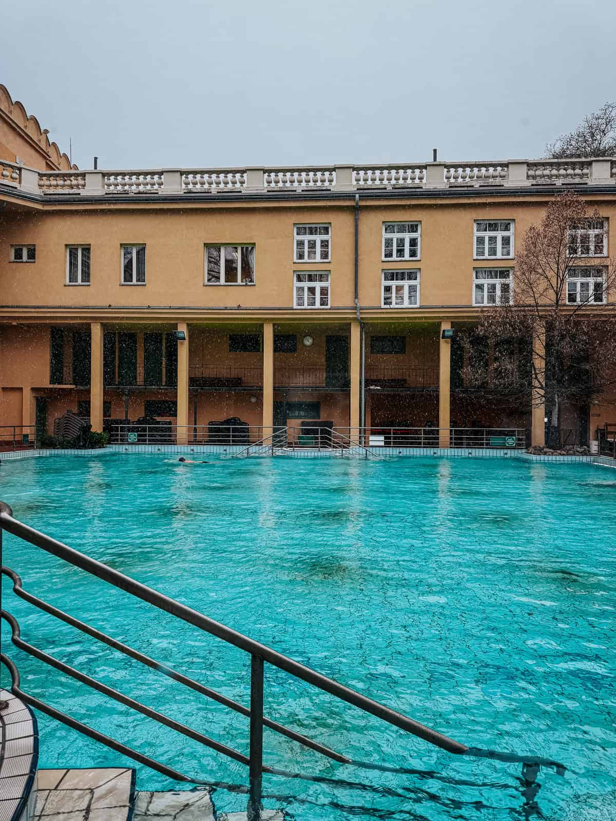 Outdoor pools at Lukács Thermal Bath with clear blue water, surrounded by yellow buildings and lifebuoys along the edges.