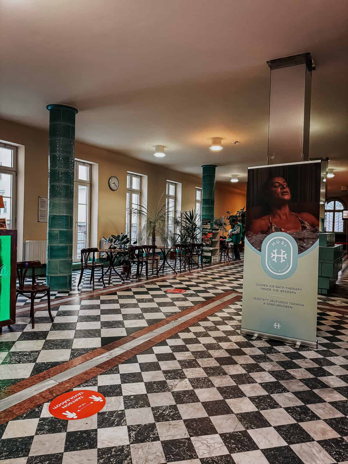 Lobby area of Lukács Thermal Bath featuring green-tiled columns, checkered flooring, tables, chairs, and a promotional banner for guided ice bath therapy.