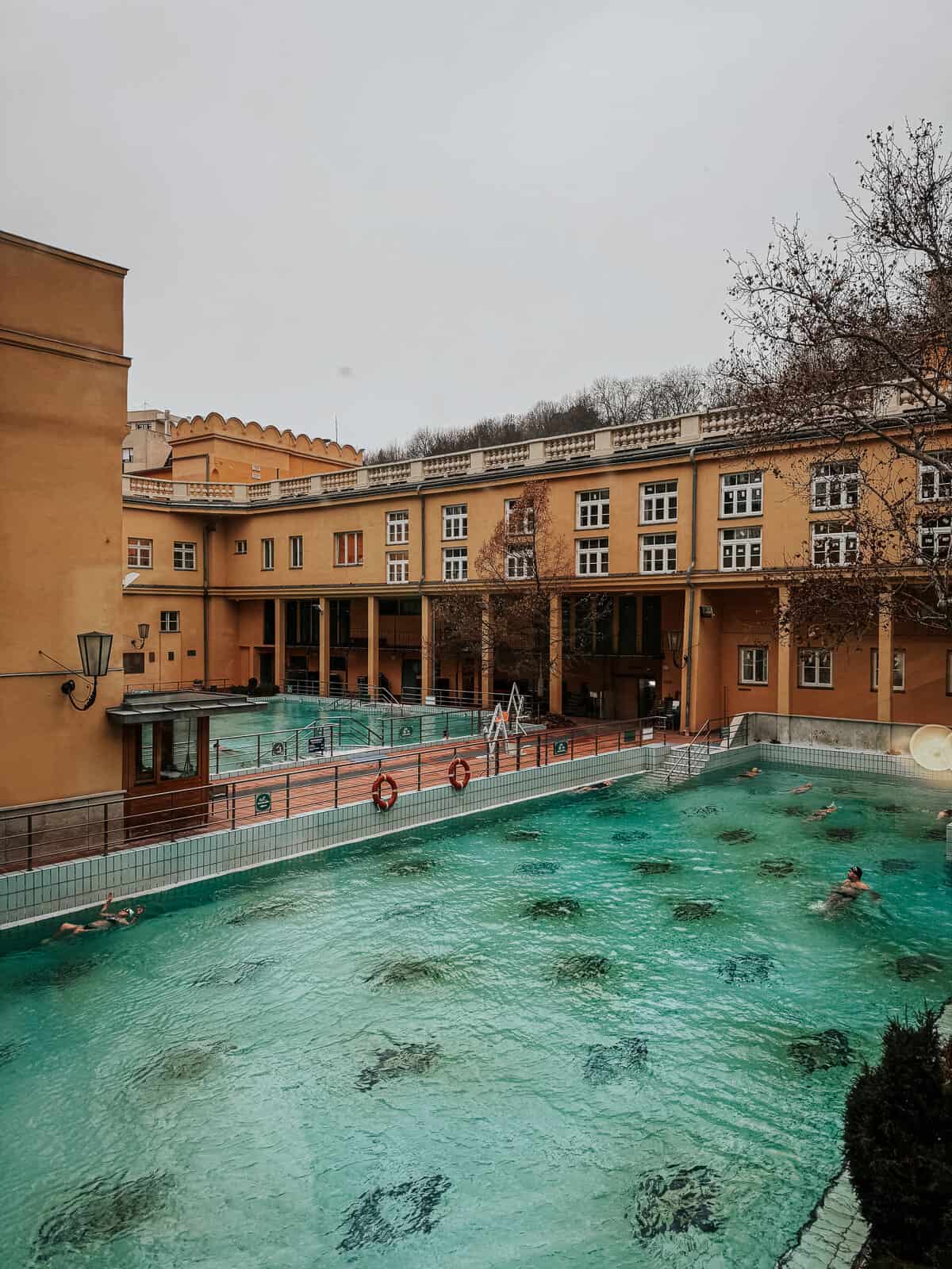 Outdoor pools at Lukács Thermal Bath with clear blue water, surrounded by yellow buildings and lifebuoys along the edges.