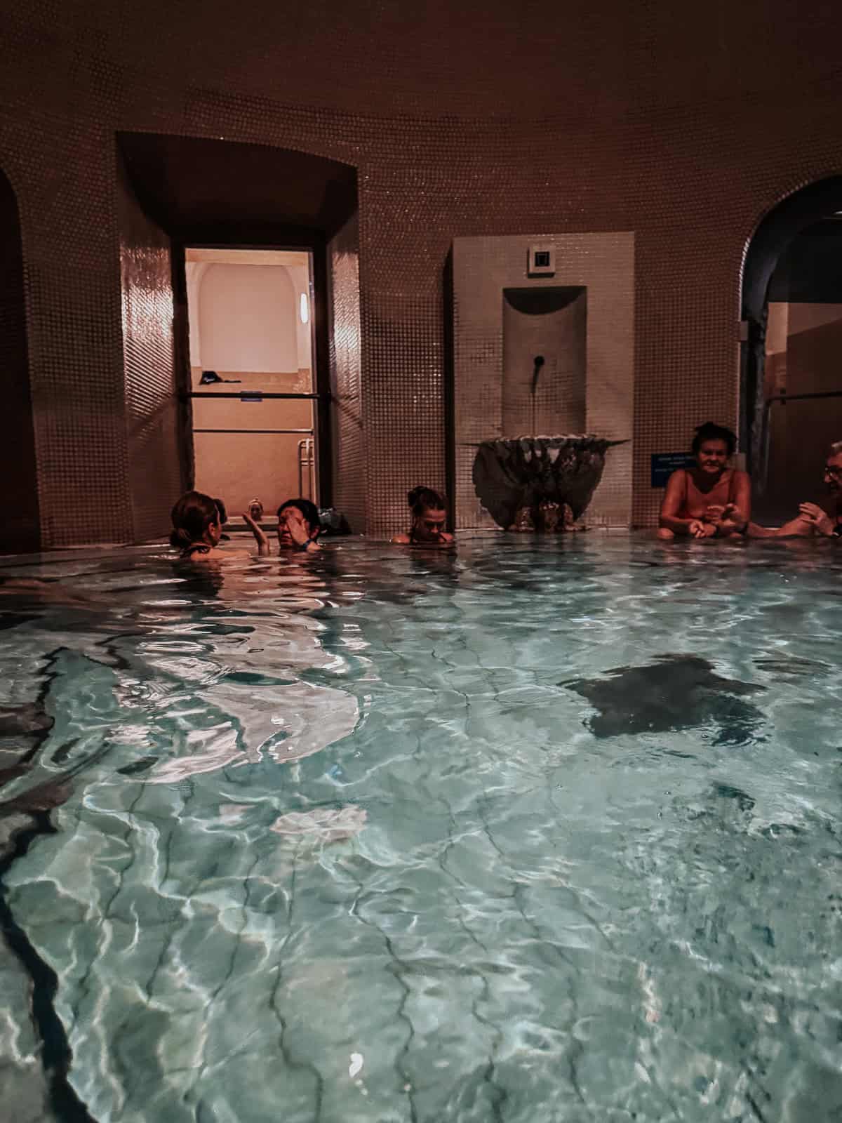 Indoor thermal pool at Lukács Thermal Bath with several people relaxing in the warm water, surrounded by mosaic-tiled walls and arched doorways.