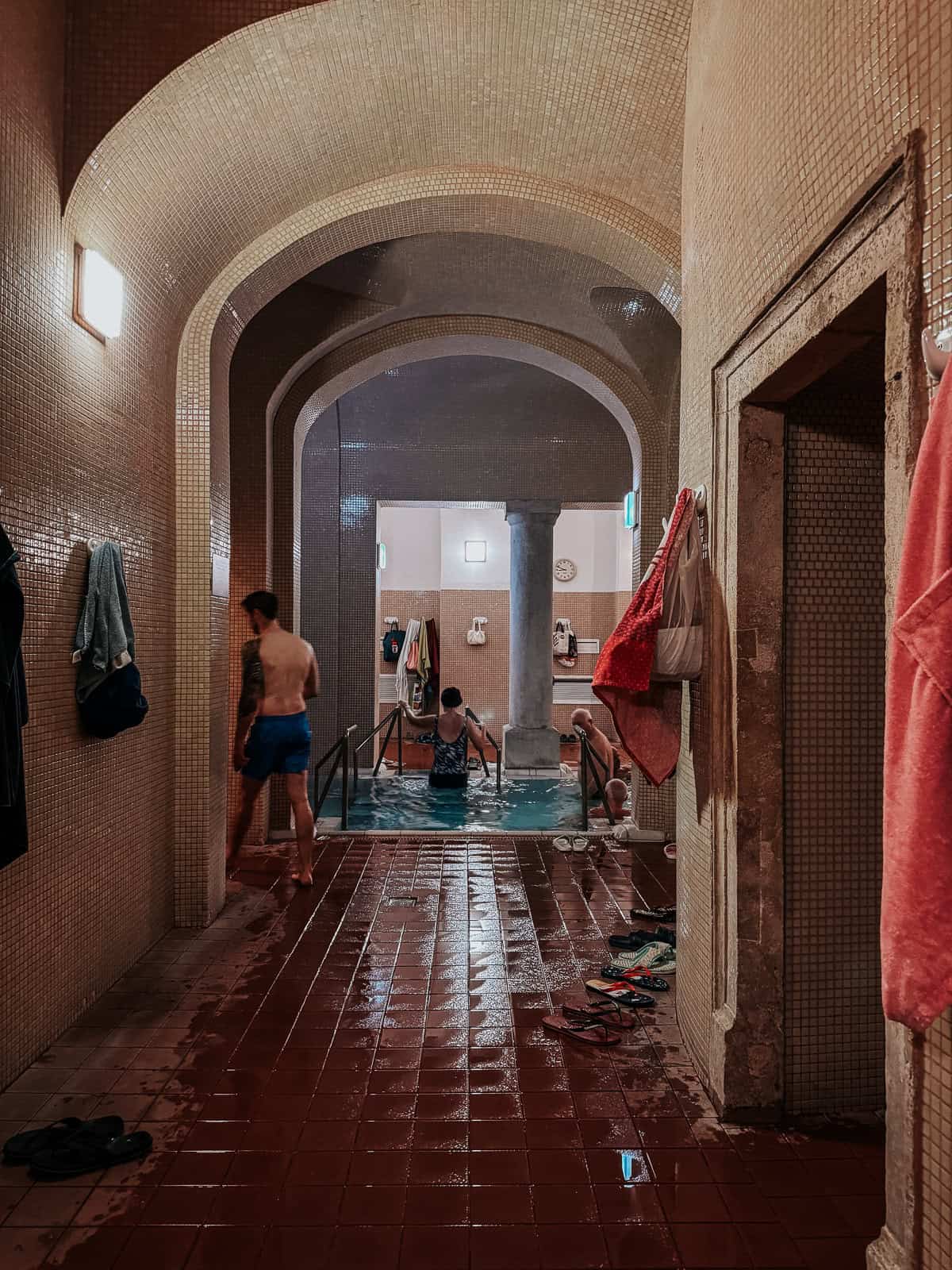 Hallway leading to an indoor thermal pool at Lukács Thermal Bath with arched ceilings, tiled walls, and people entering the pool area.