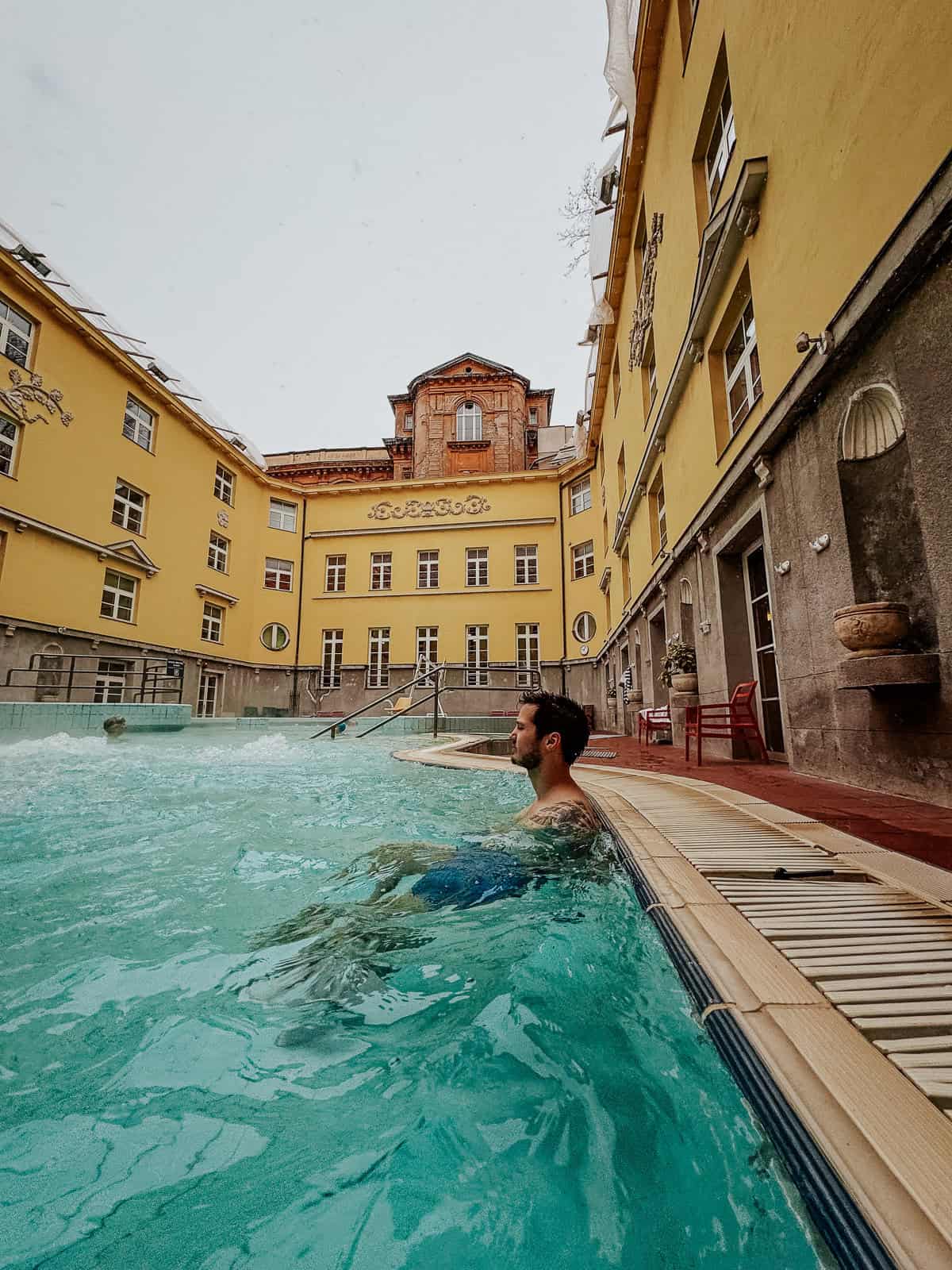 A woman in a black swimsuit stands in a turquoise thermal pool at Lukács Thermal Bath, with a yellow building in the background featuring decorative details and windows.