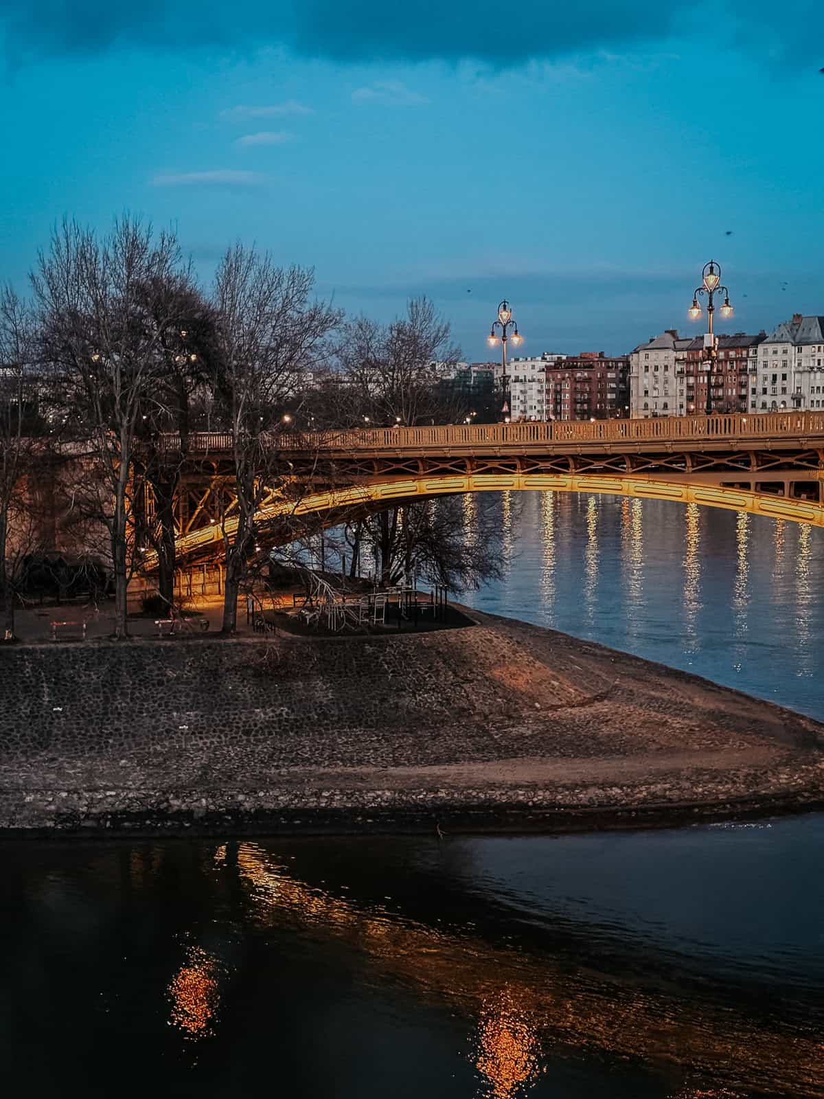 A bridge illuminated with warm lights at dusk, reflecting in the river below with buildings in the background on Margaret Island.