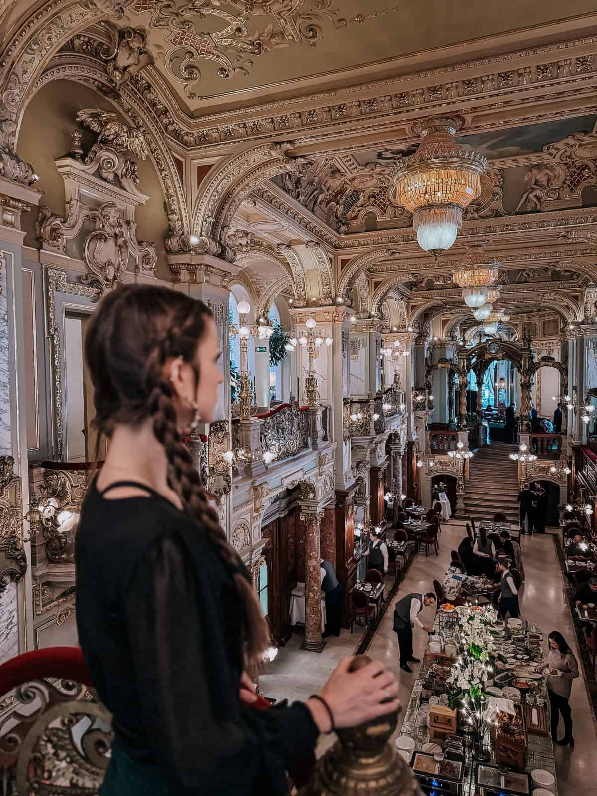 A woman with braided hair stands on a balcony, overlooking a grand, ornate hall with chandeliers and intricate architectural details. Below, waitstaff and guests are seen at dining tables in an elegant, historic setting.