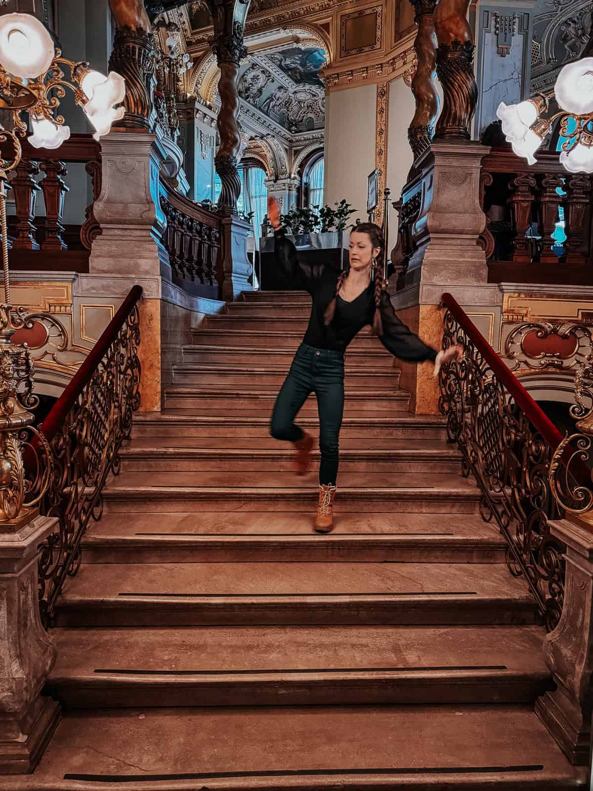 A woman with braided hair is captured mid-motion on a grand staircase, with ornate railings and opulent decor surrounding her. The background features lavish architectural details and soft lighting from decorative lamps.