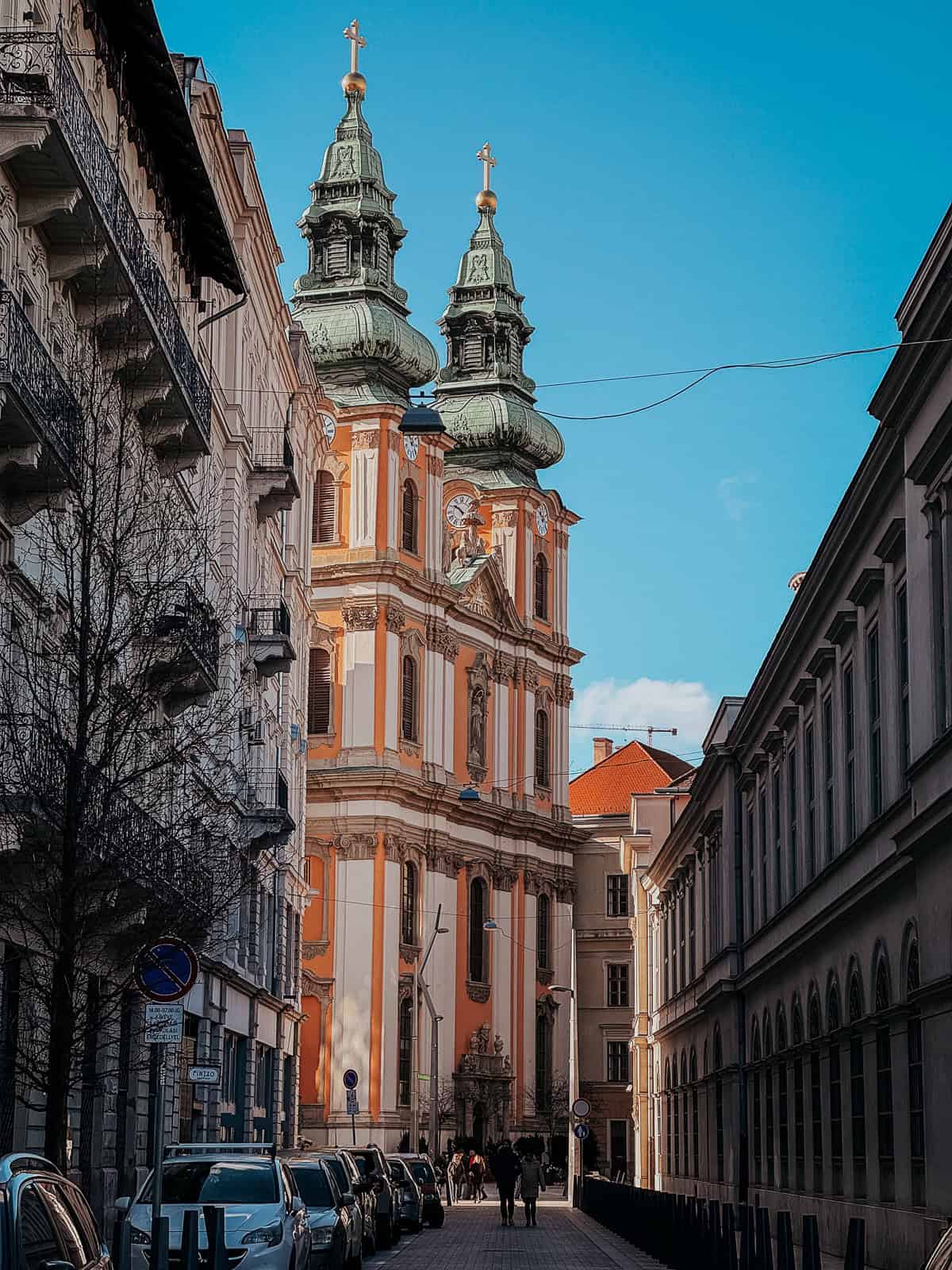 A street view of a baroque-style church with twin green domes in Budapest, framed by adjacent buildings