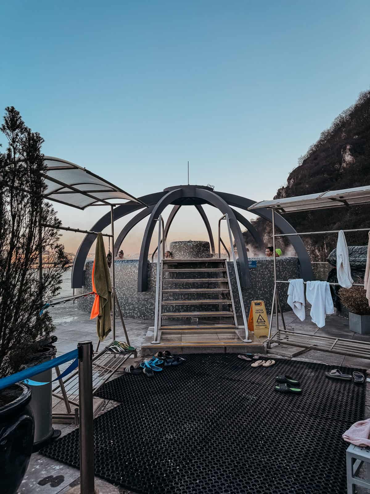 The rooftop of Rudas Baths, showcasing a modern structure with a small pool area. Various towels and sandals are scattered around, and a few people are visible in the background, enjoying the view.