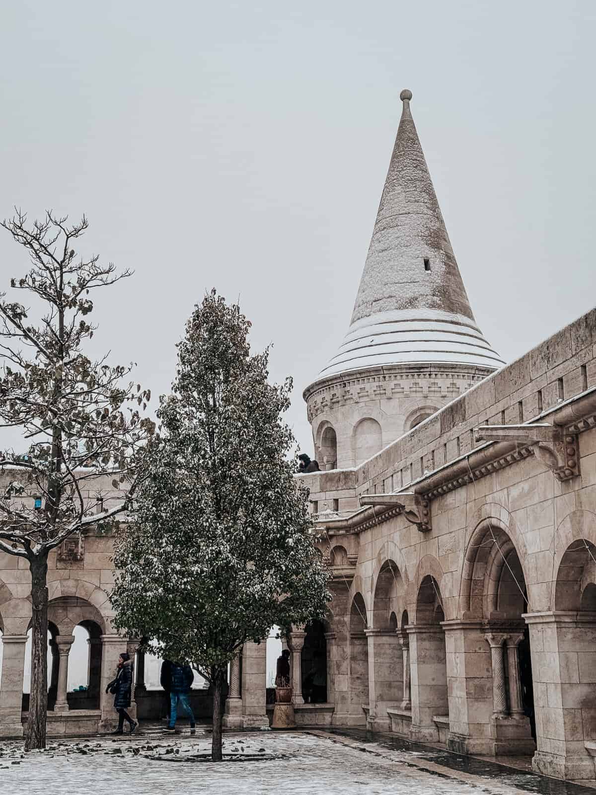 A snowy day at Fisherman's Bastion in Budapest, featuring an ornate stone tower with a conical roof, framed by a courtyard with arched walkways and a tree in the foreground.