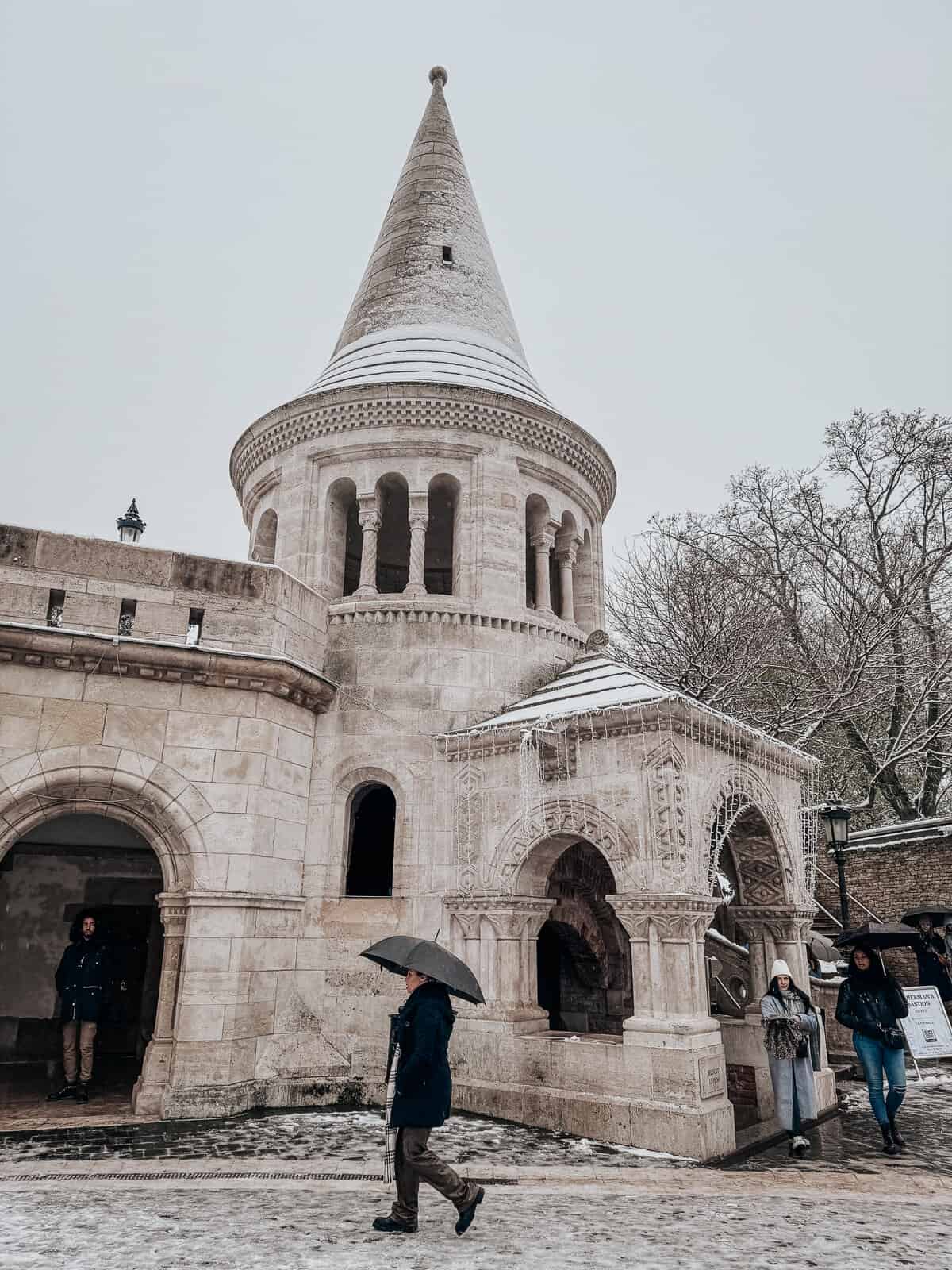 Visitors walking under umbrellas on a snowy day at Fisherman's Bastion, with a prominent stone tower and intricate arched entrances