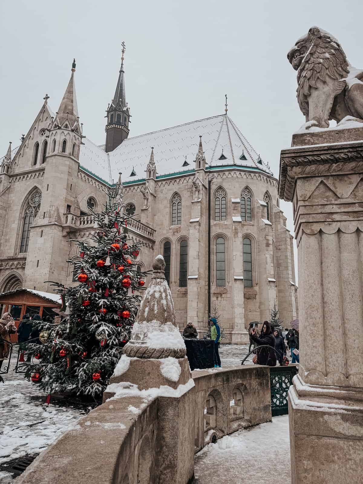 The exterior of Fisherman's Bastion decorated with Christmas ornaments on a tree, showcasing Gothic architectural details and a statue of a lion in the foreground.