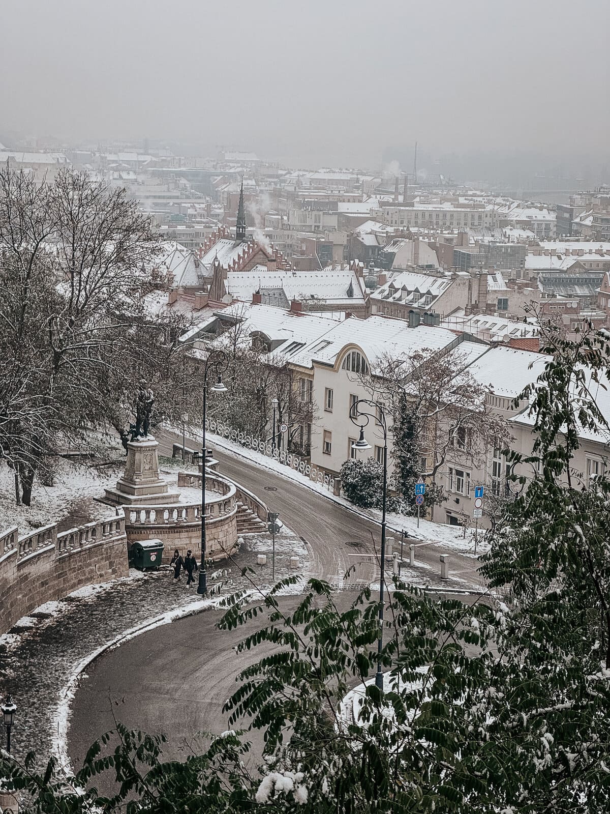 Snow-covered cityscape of Budapest viewed from Fisherman's Bastion, showing rooftops, a winding road, and a distant skyline shrouded in mist.