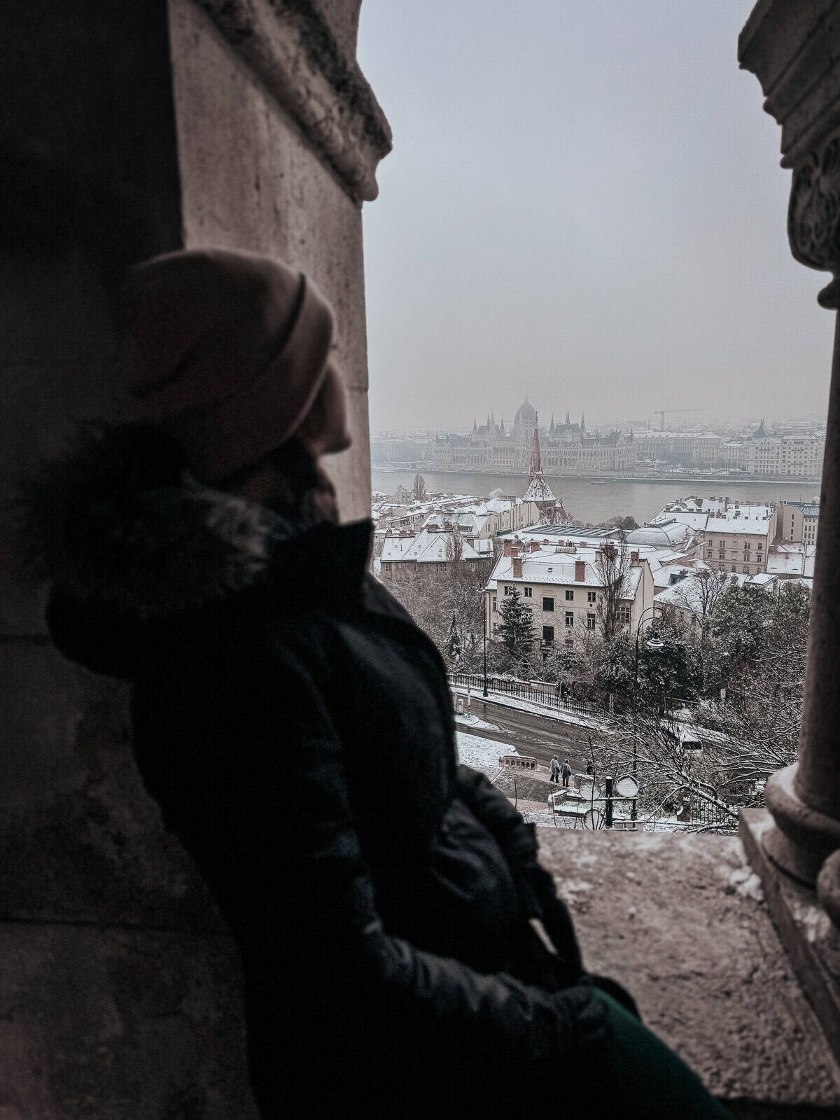 A person in winter clothing looking out from Fisherman's Bastion over a snowy Budapest cityscape, with the Danube River and iconic buildings in the background.