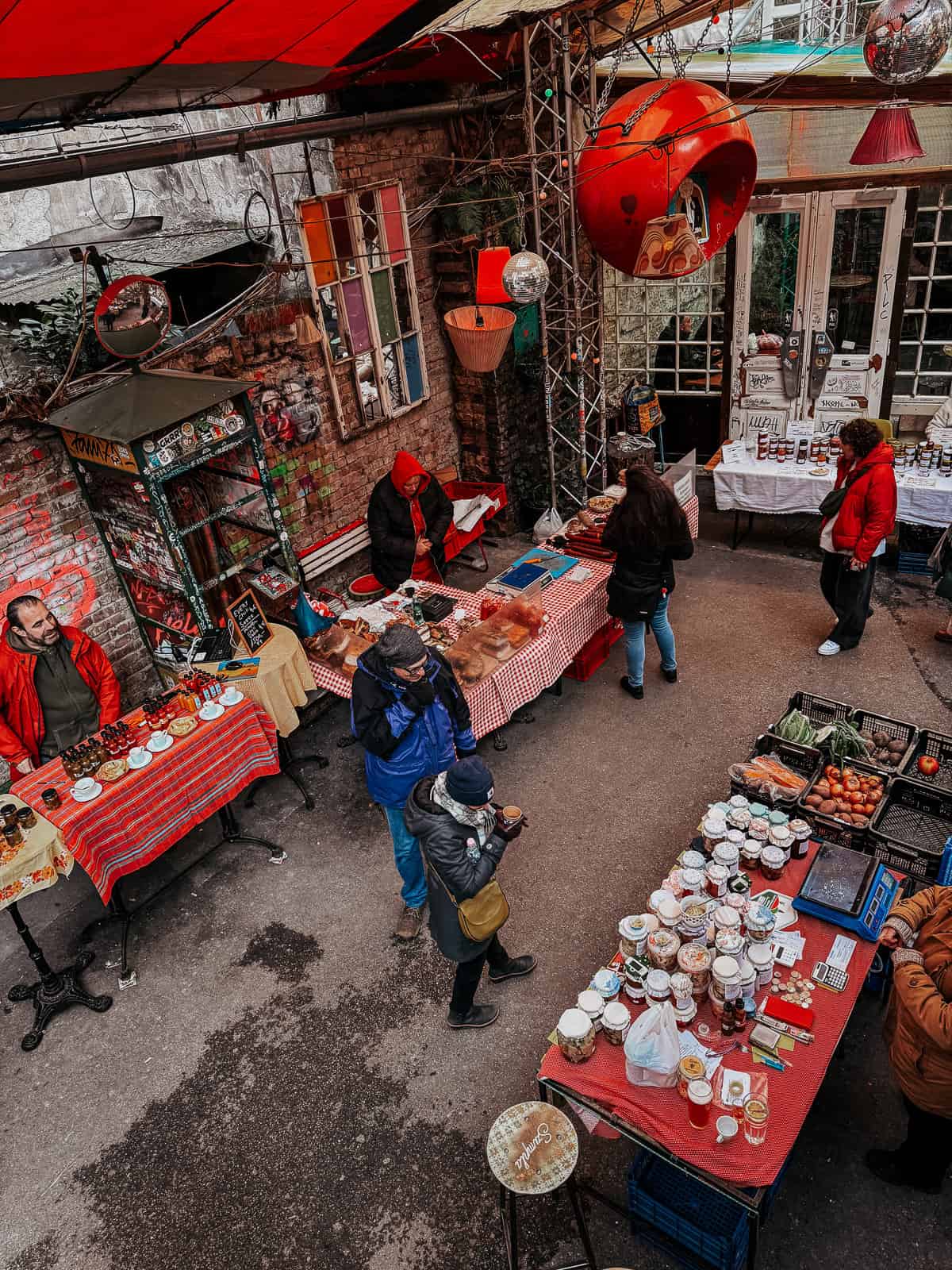 An overhead view of a ruin bar farmers market with various vendors selling their goods at tables covered with red checkered cloths. People are seen browsing and engaging with the vendors in a vibrant, eclectic setting