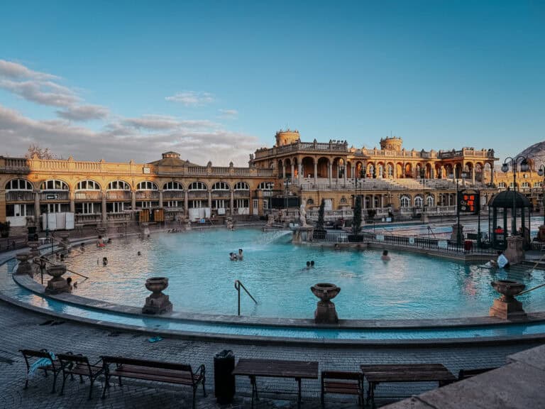 The grand outdoor pool at Szechenyi Baths, with people swimming and relaxing in the thermal waters. The historic bathhouse building surrounds the pool, featuring arches and decorative elements under a bright blue sky.