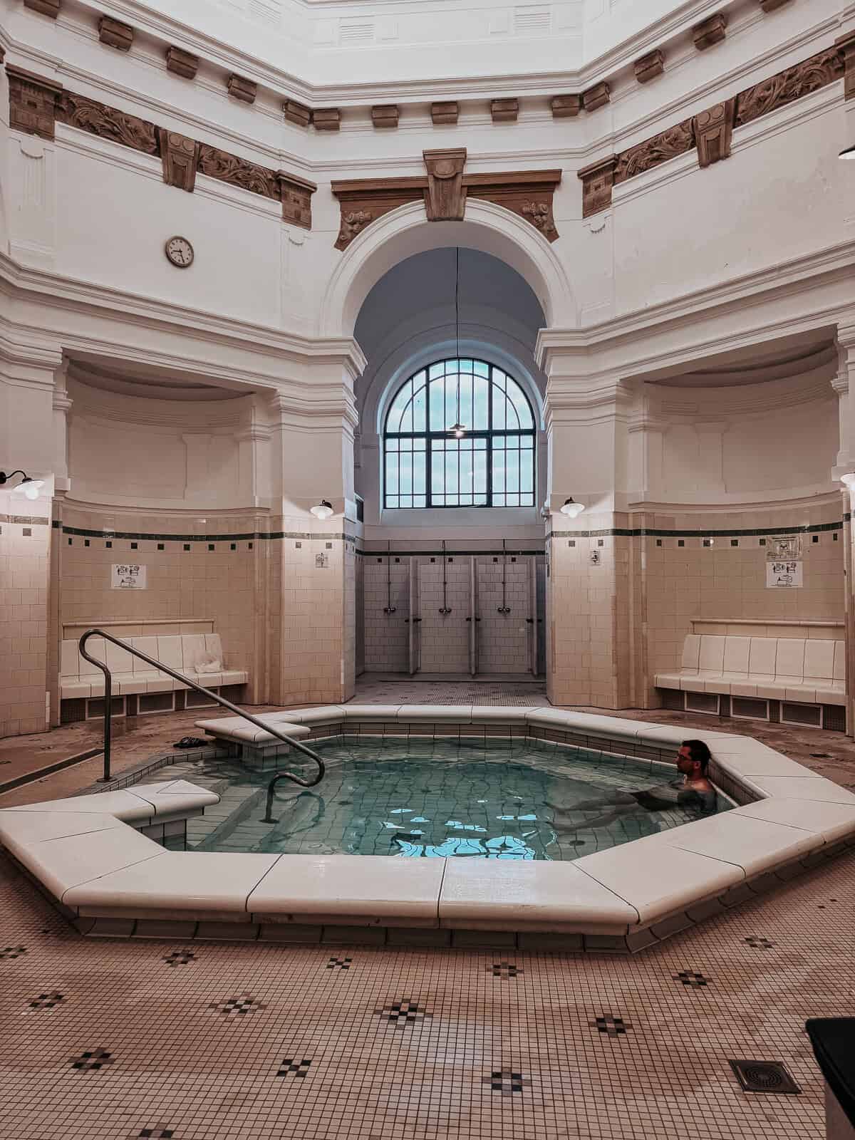 An indoor pool at Szechenyi Baths, with a lone person soaking in the warm water. The high, white walls and arched windows create a spacious and elegant atmosphere.