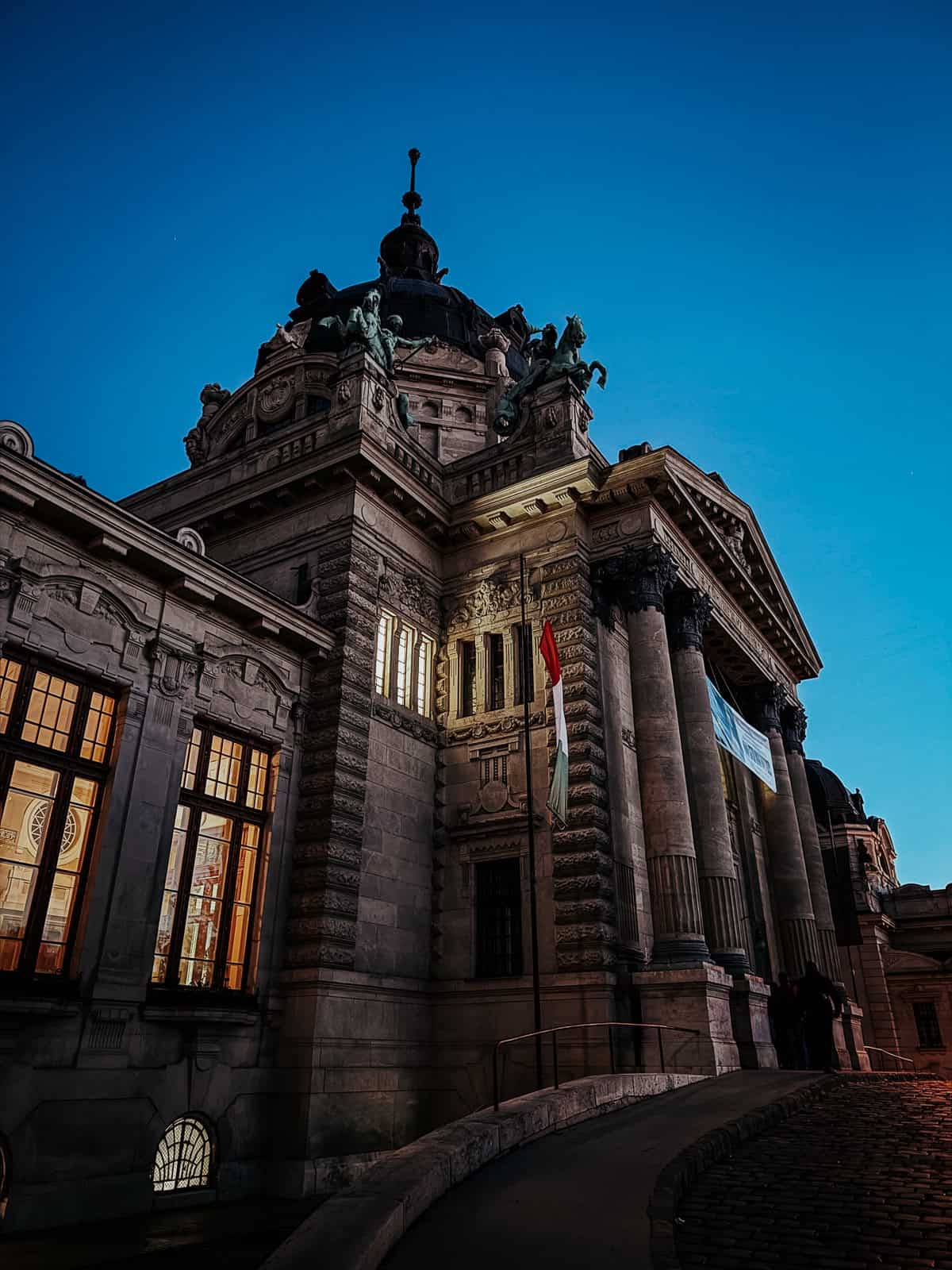 A grand, historic building with a dome and statues on top, illuminated against a twilight sky. The architectural details are highlighted by the evening light.