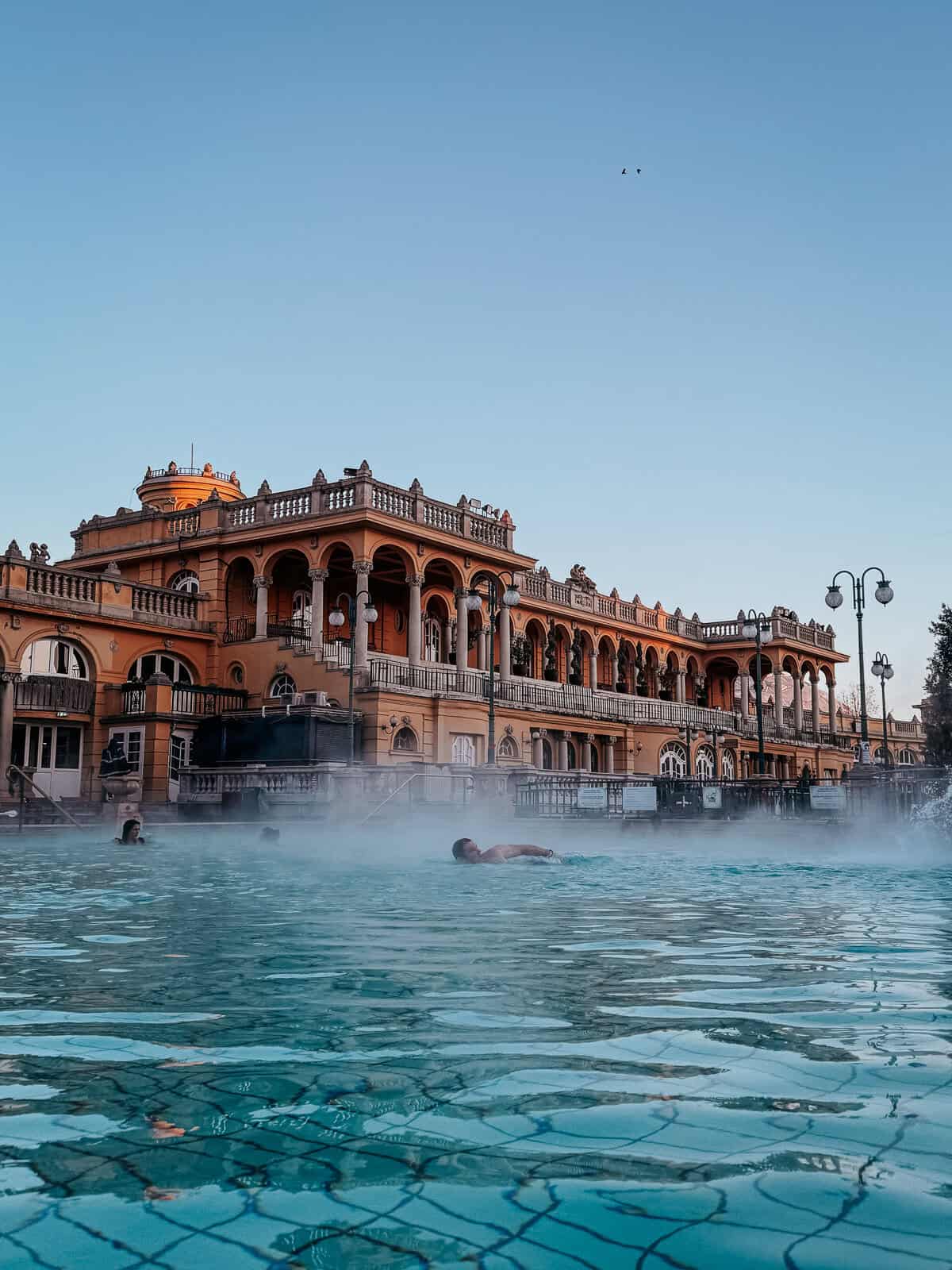 The main outdoor pool with people swimming, surrounded by grand, ornate buildings under a clear sky, with steam creating a mystical atmosphere.