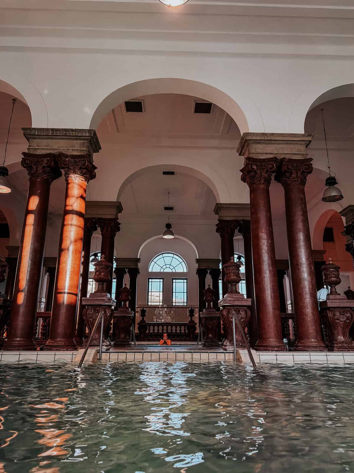 An indoor pool area with grand columns, arches, and warm lighting reflecting off the water, emphasizing the luxurious and historic architecture.