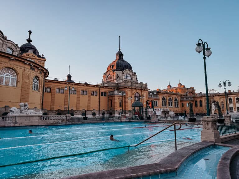 An outdoor thermal pool with steam rising from the water, surrounded by the historic building. The pool is lit by warm lights, creating a cozy ambiance during twilight.