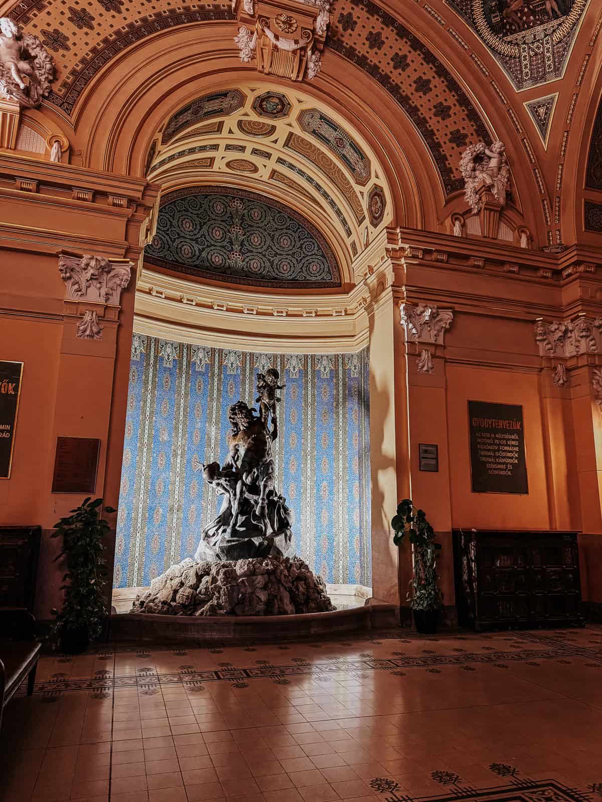 A lavish interior featuring a detailed, ornate ceiling and a central fountain with statues, surrounded by decorative tiles and rich patterns.