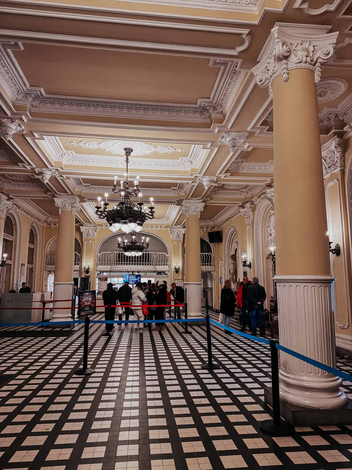 The lobby area with classic architectural elements, including tall columns, a chandelier, and a checkered floor. People are waiting in line, adding to the lively atmosphere.