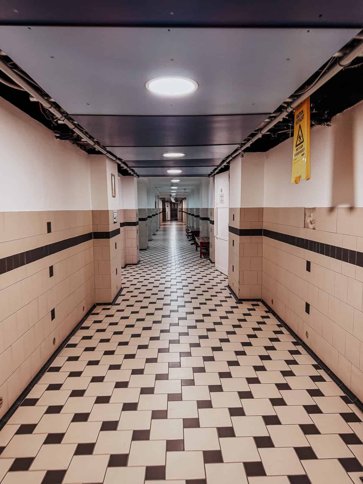 A long, tiled hallway with a checkered pattern floor, lit by ceiling lights. The walls are lined with white tiles and occasional signs.