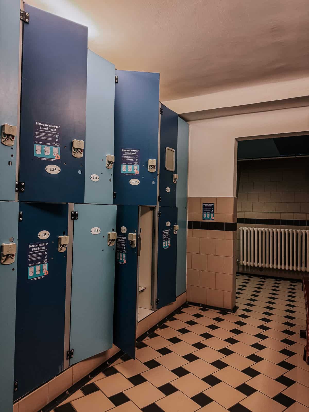 A locker room with blue lockers, some open, and instructional signs on the doors. The area is clean and well-maintained with a checkered tile floor.