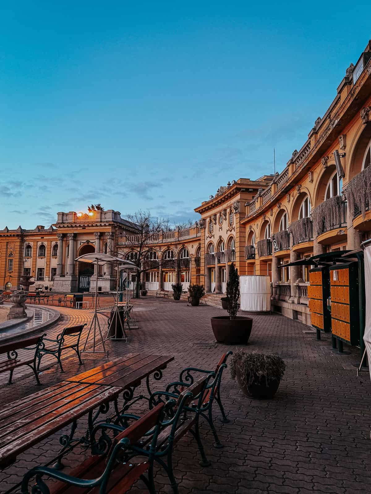 A view of the courtyard with benches and tables, surrounded by the historic architecture of the thermal baths. The area is empty, providing a serene atmosphere.