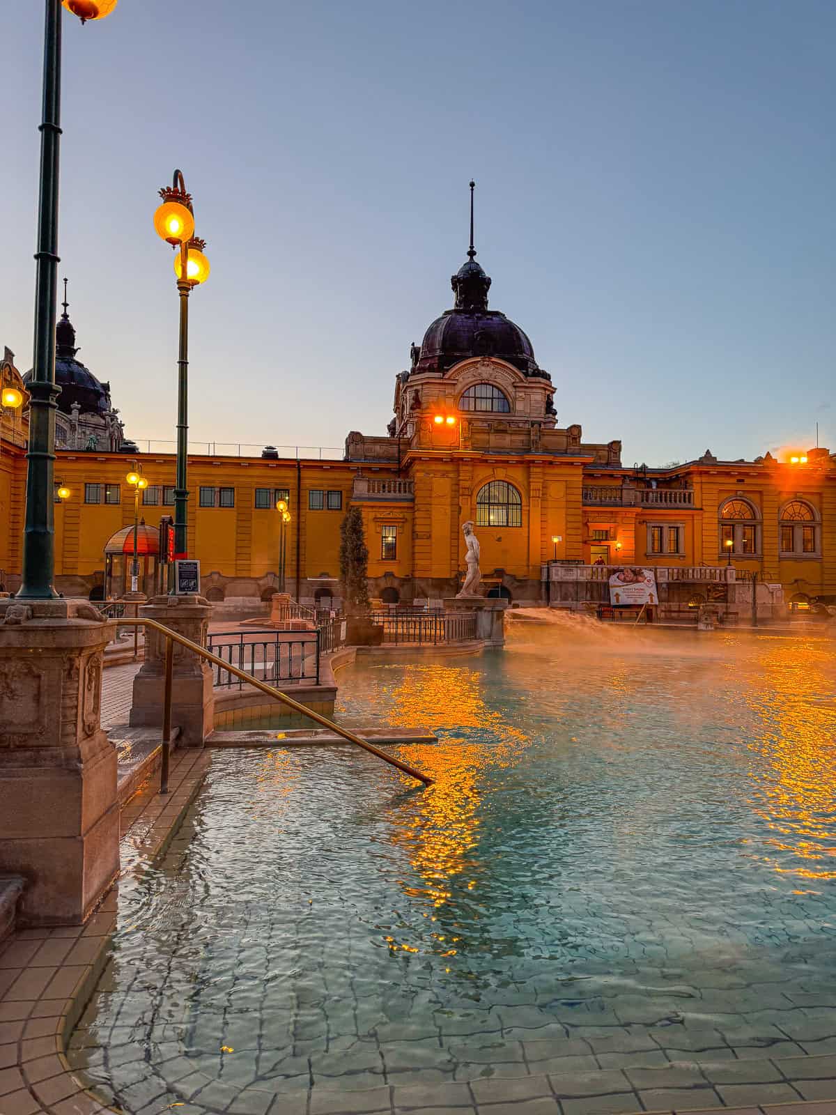 An outdoor pool with steam rising, surrounded by the illuminated historic building with dome structures and lit lamp posts.