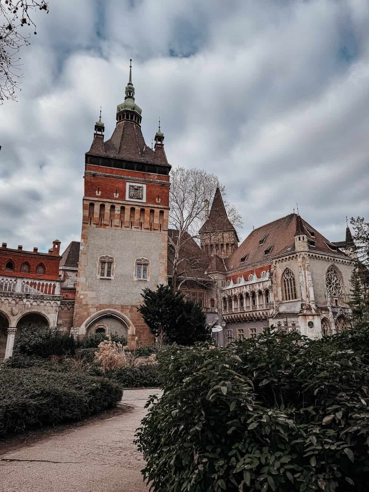 A view of Vajdahunyad Castle in Budapest, featuring its eclectic architectural styles and lush garden under a cloudy sky.