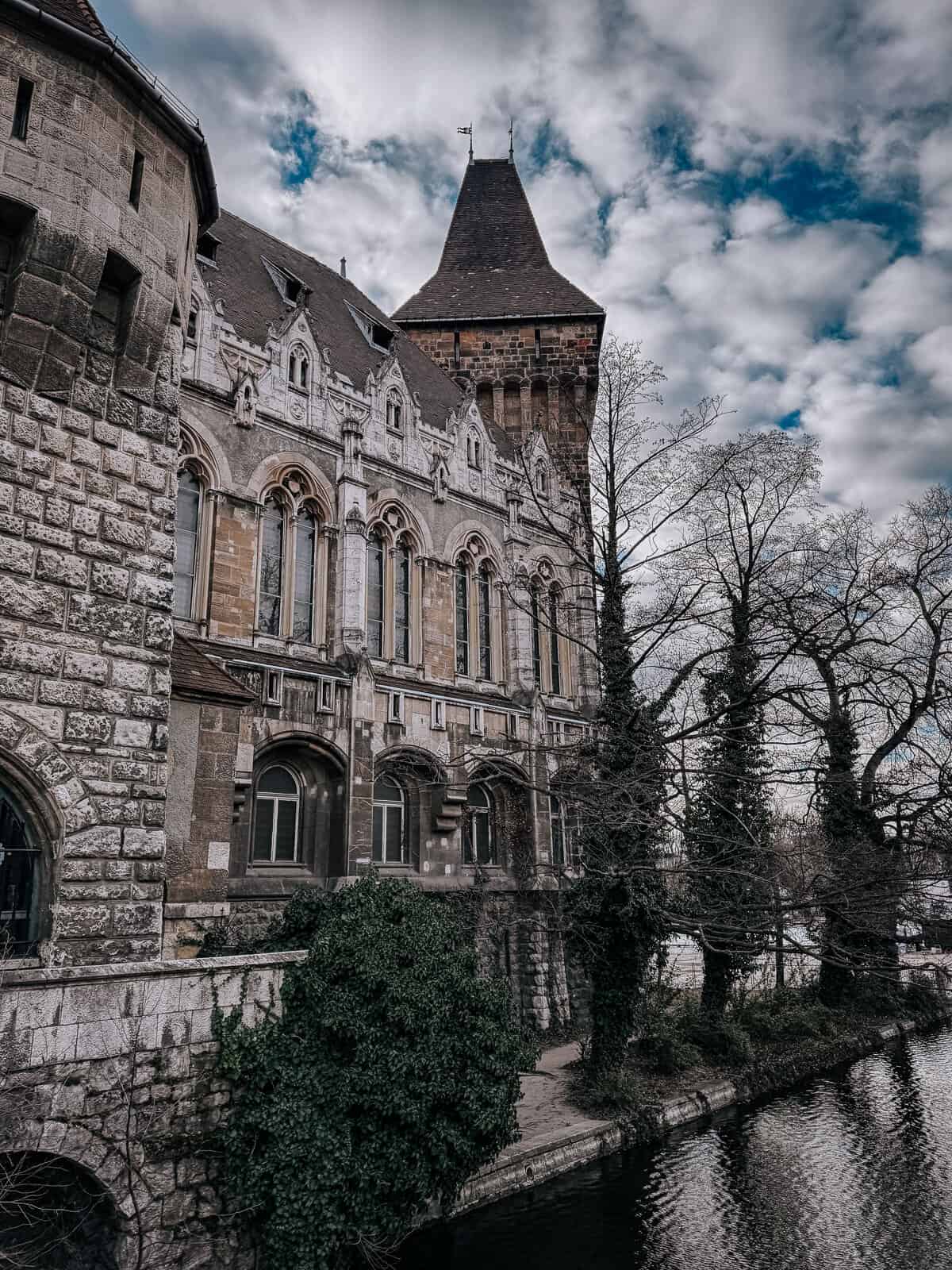 A picturesque view of Vajdahunyad Castle's ornate facade and tower against a backdrop of cloudy sky. The castle is partially covered with ivy, and a river runs alongside the building.