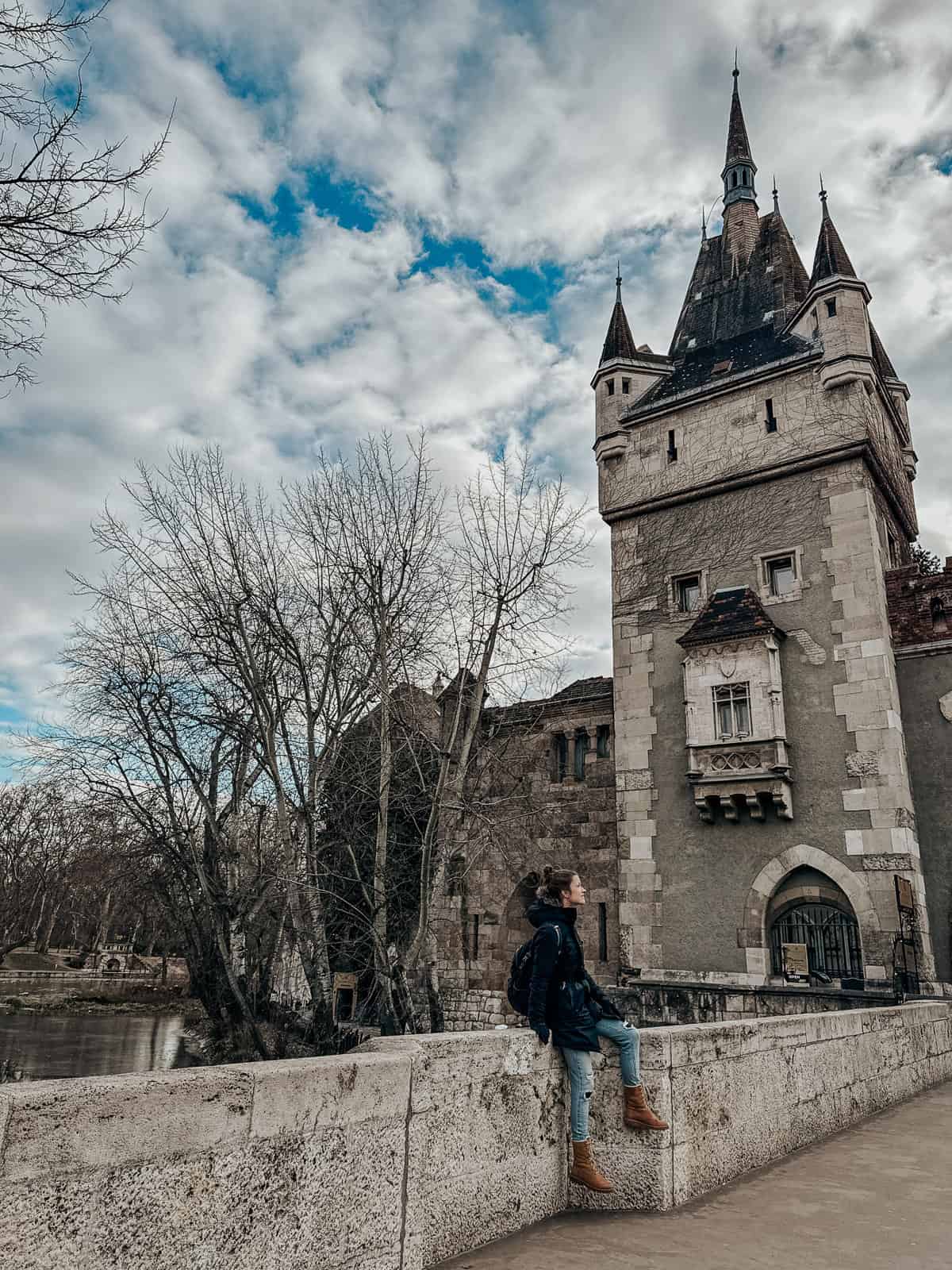 A person in a black coat and boots leans against the stone railing of a bridge, with the spires of Vajdahunyad Castle towering in the background under a partly cloudy sky.