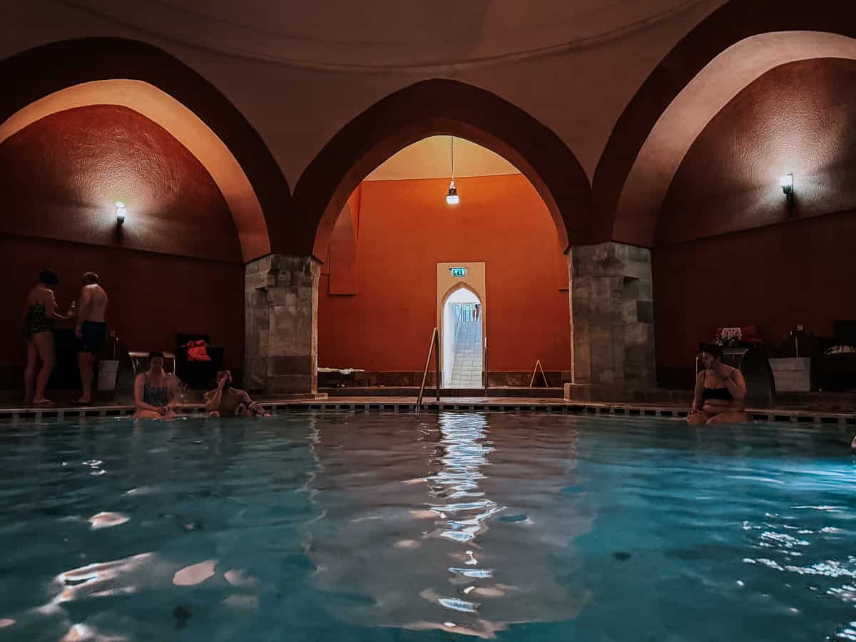 The interior of Veli Bej Baths, showing a tranquil pool area with arches and a soft, warm light. A few people are sitting by the pool, enjoying the peaceful ambiance.