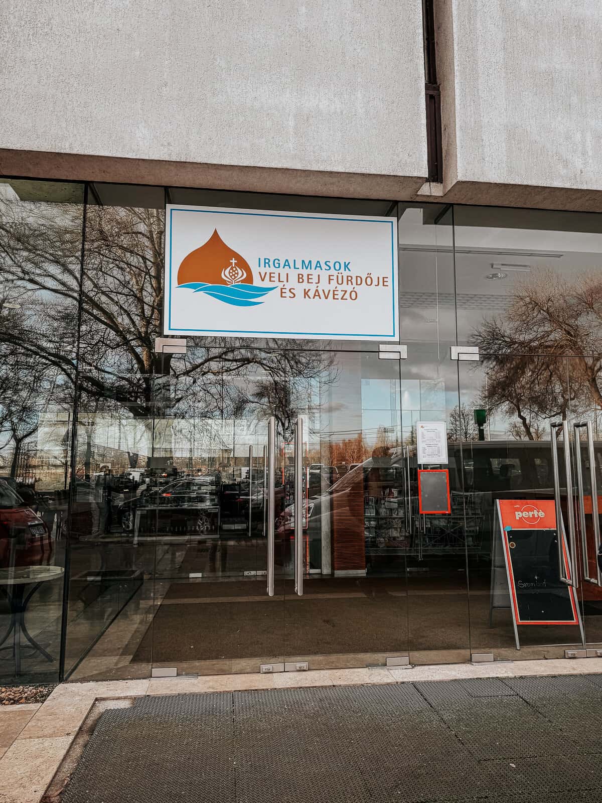 The entrance of Irgalmasok Veli Bej Fürdője és Kávézó, a glass-fronted building with a sign displaying the name above the door, reflecting the street and trees outside.