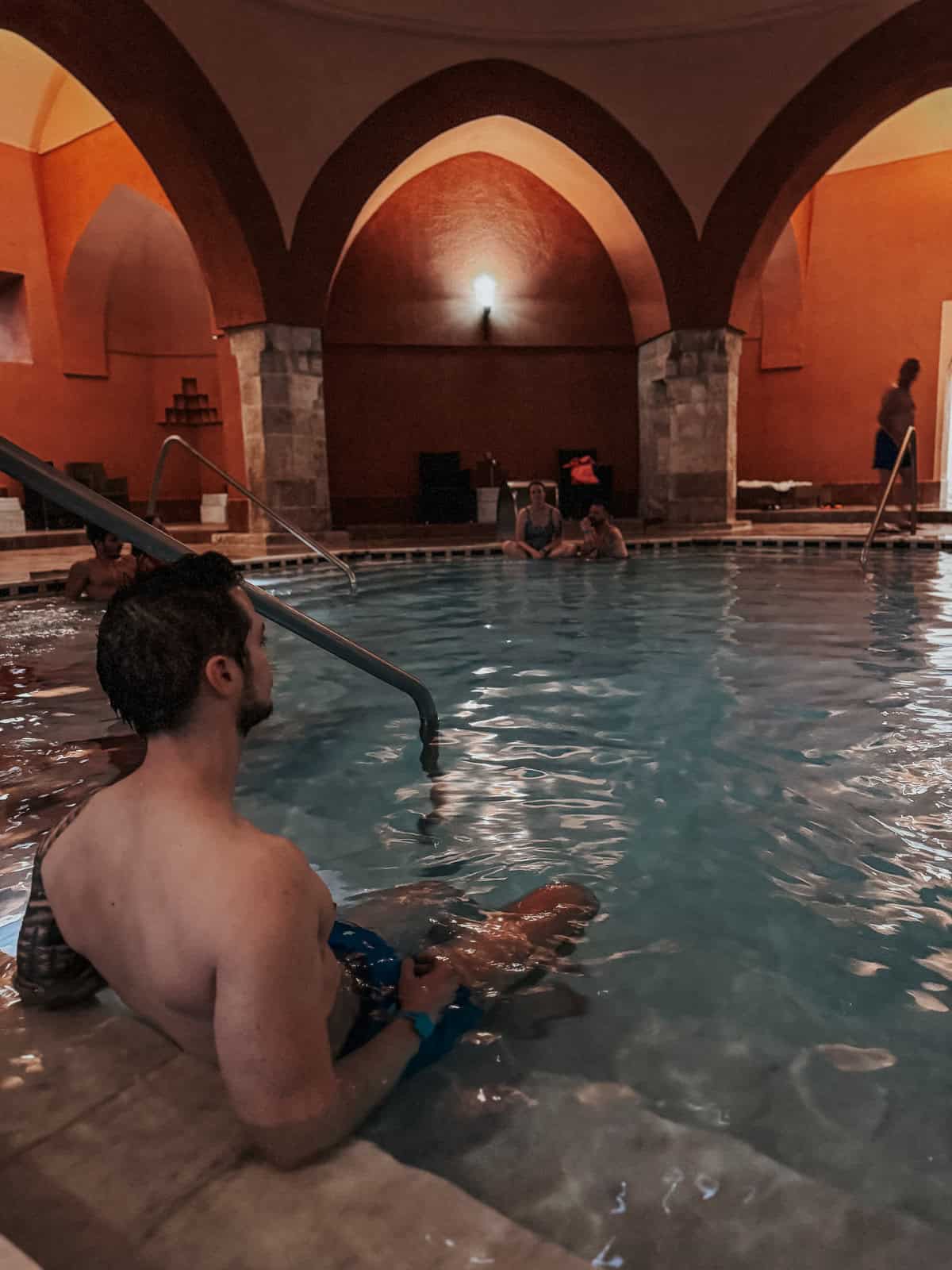 A man sitting by the edge of the indoor pool, resting his arms on his knees, with other bathers visible in the background under arched ceilings.