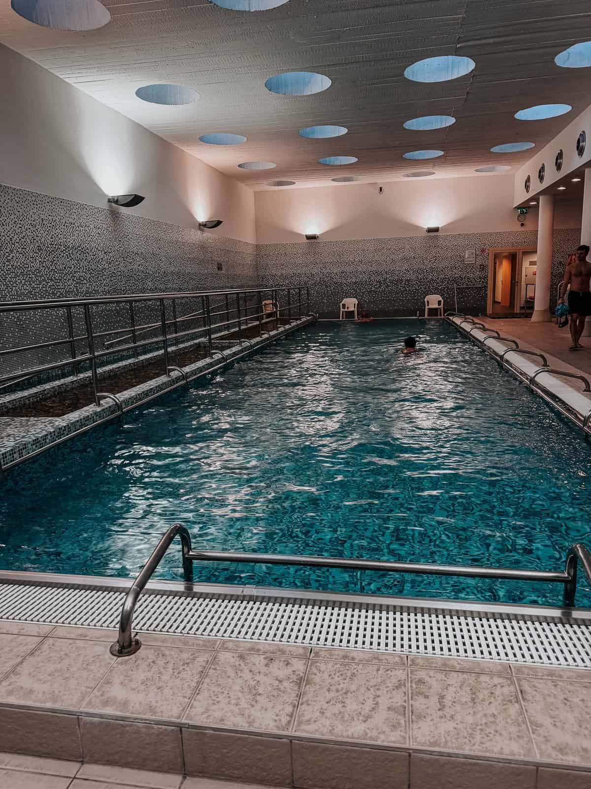 A larger, rectangular swimming pool with a modern design, featuring a tiled floor and ceiling lights creating a pattern of circular light spots.