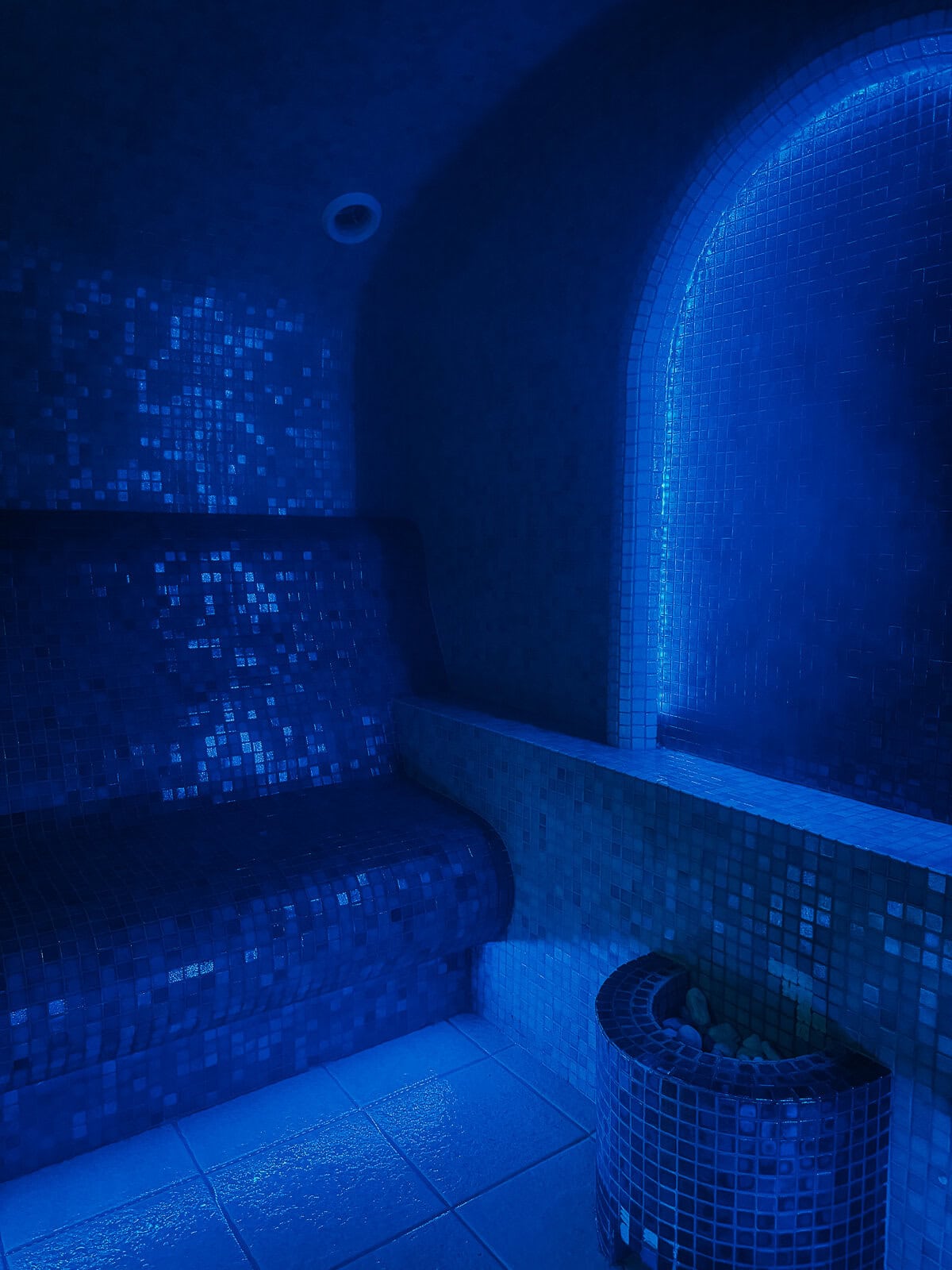 A dimly lit steam room with blue-tiled seating and a serene, calming blue light ambiance