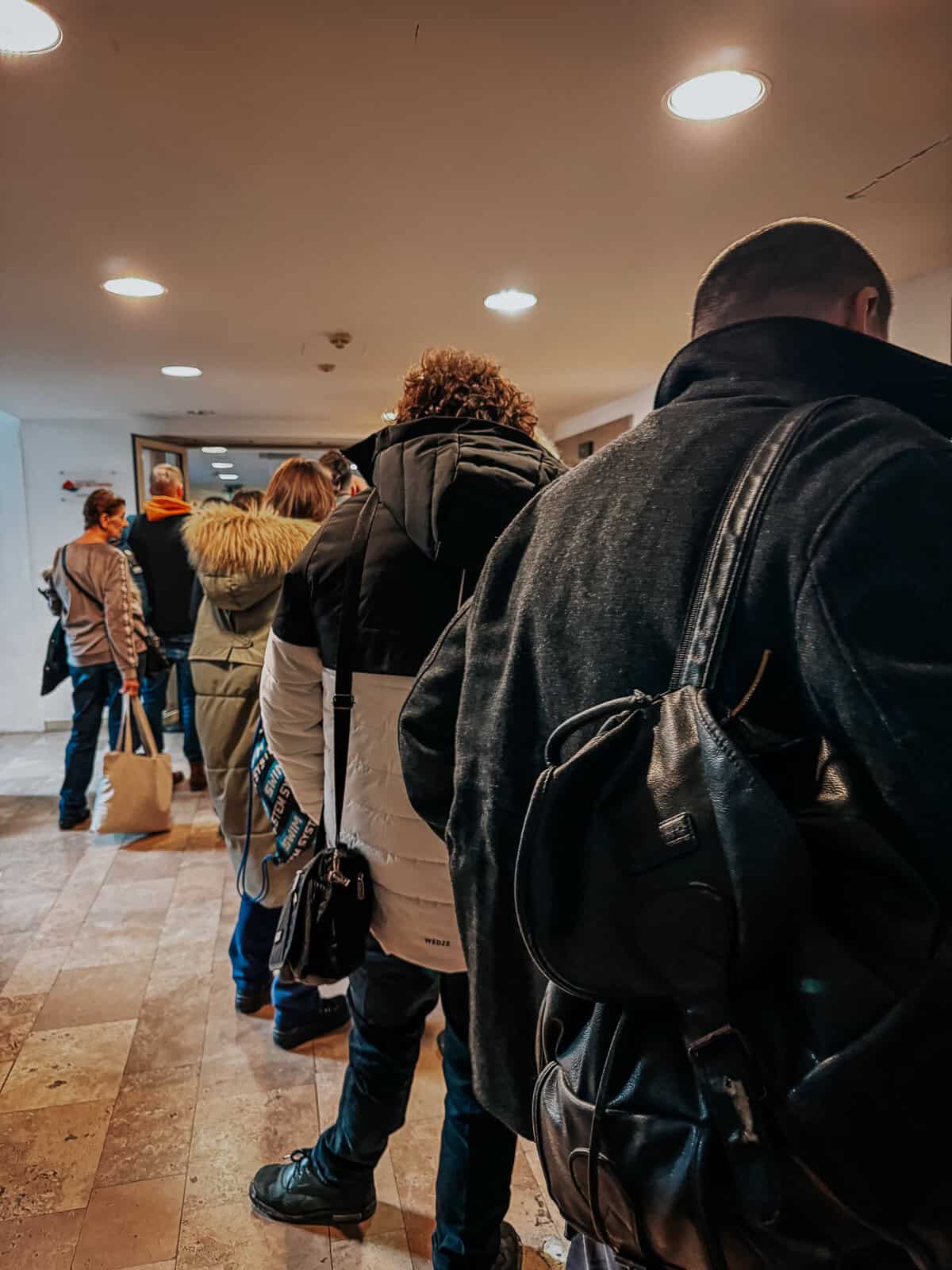 A queue of people in winter clothing waiting inside a corridor, facing forward and standing under bright ceiling lights.