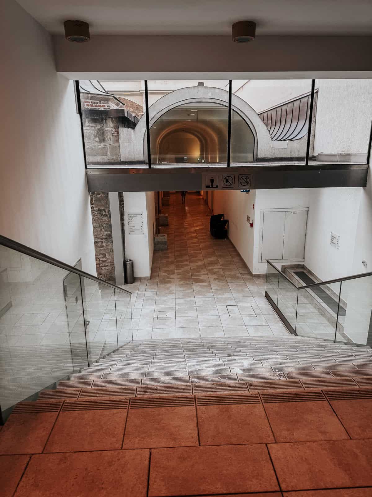 A stairway leading down to a hallway with tiled floors and modern glass railings, illuminated by natural light from large windows above.