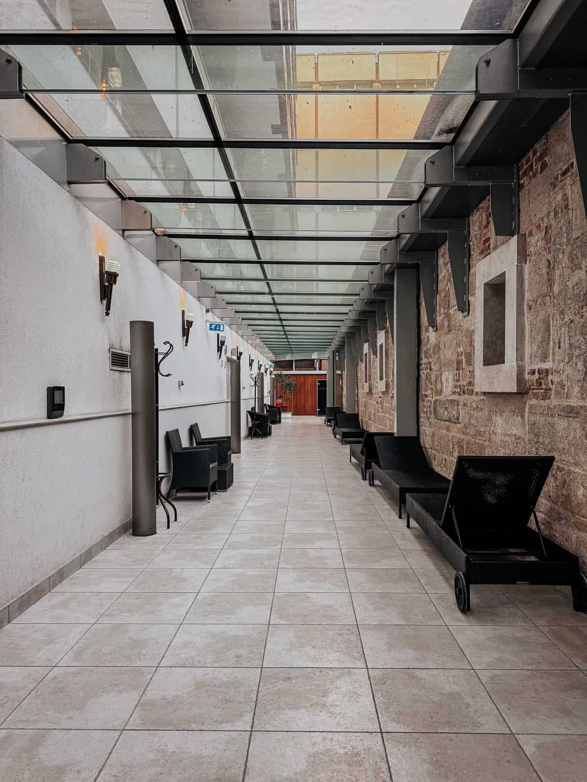 A long, modern hallway with a glass ceiling and lounge chairs along the sides, featuring a blend of contemporary and historical architecture.