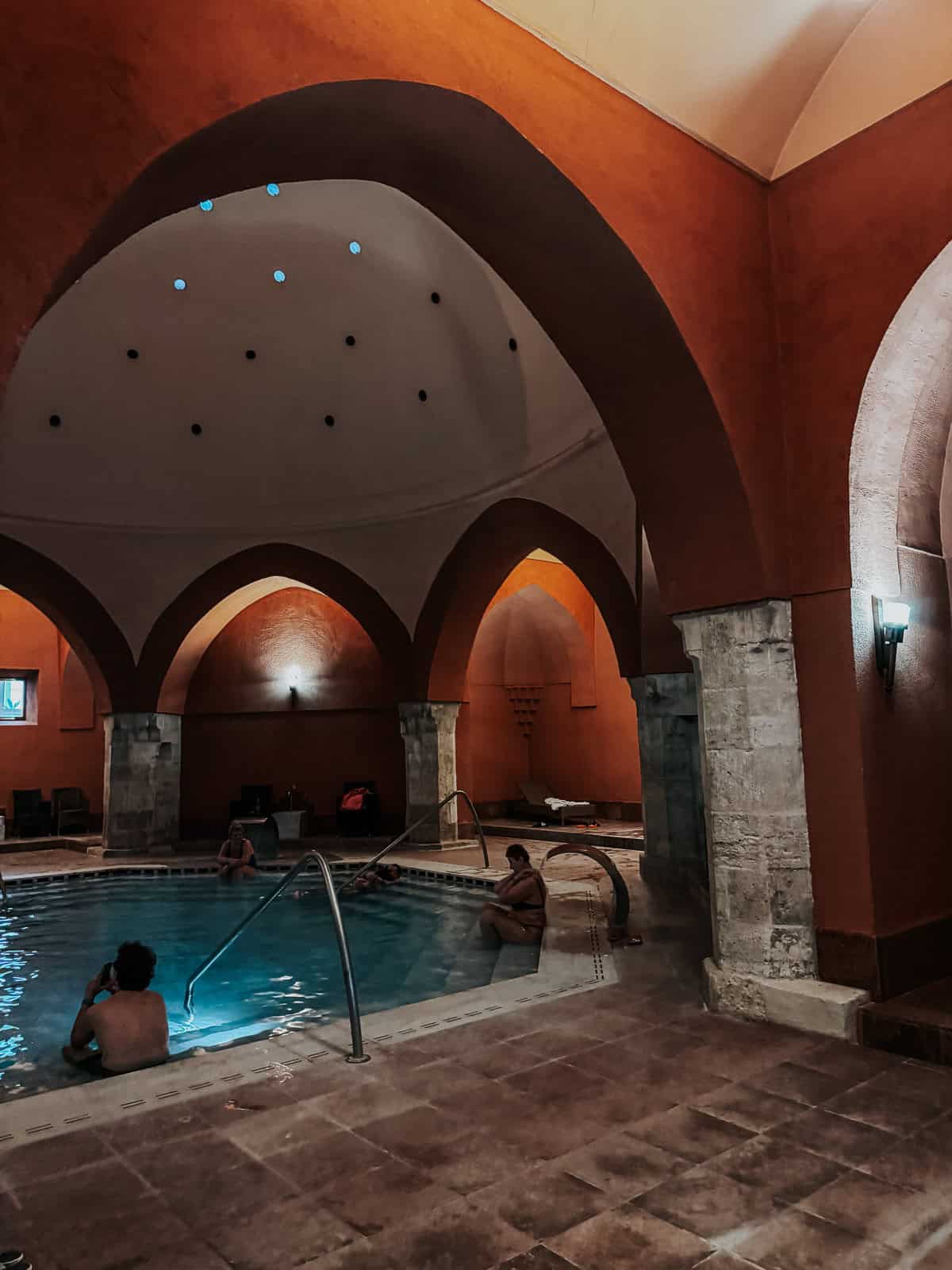 An indoor pool with people relaxing in the water, surrounded by arches and dim lighting, creating a serene ambiance.