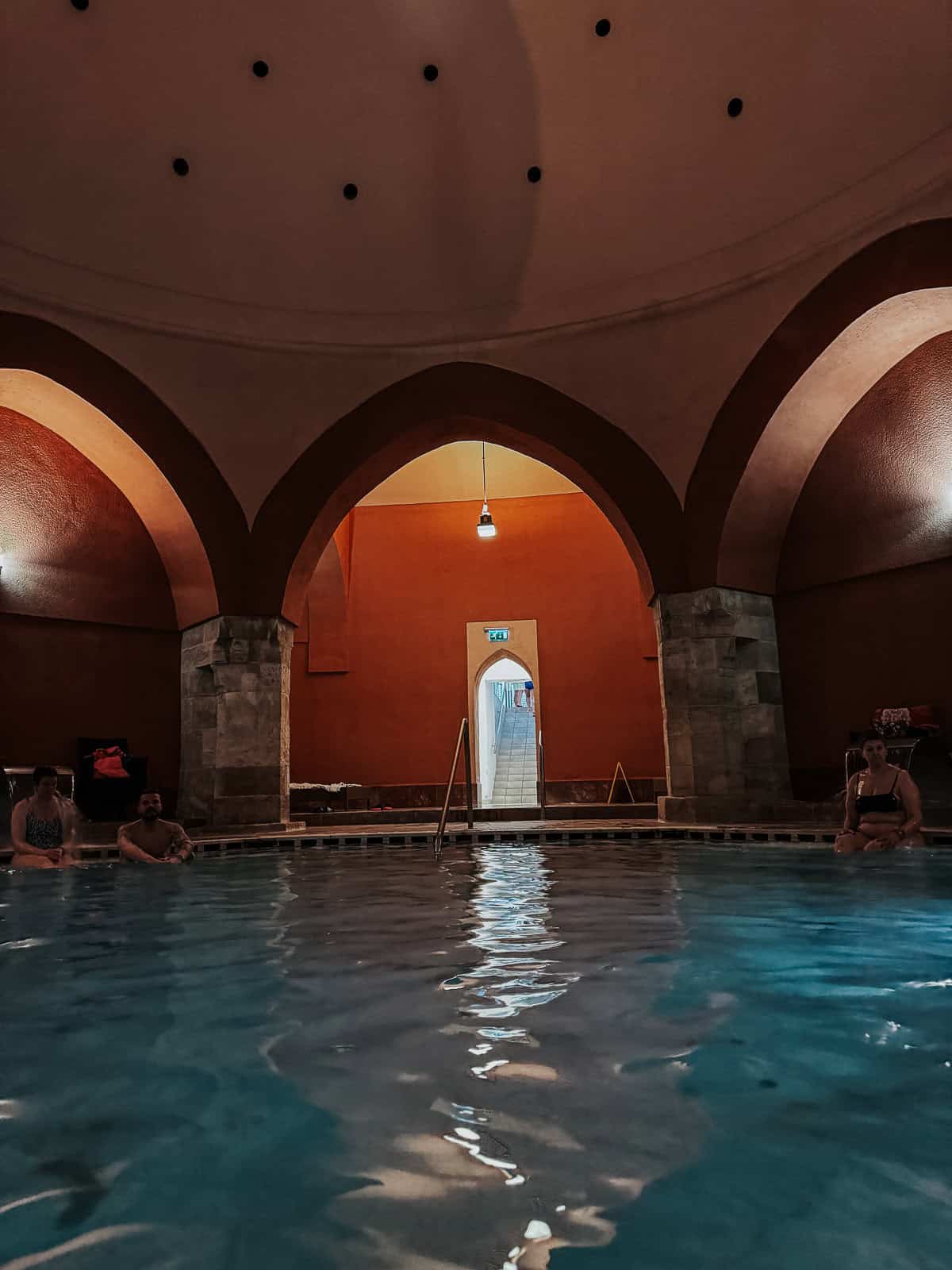 wider shot of the indoor pool area, emphasizing the vaulted ceilings, warm lighting, and people enjoying the water