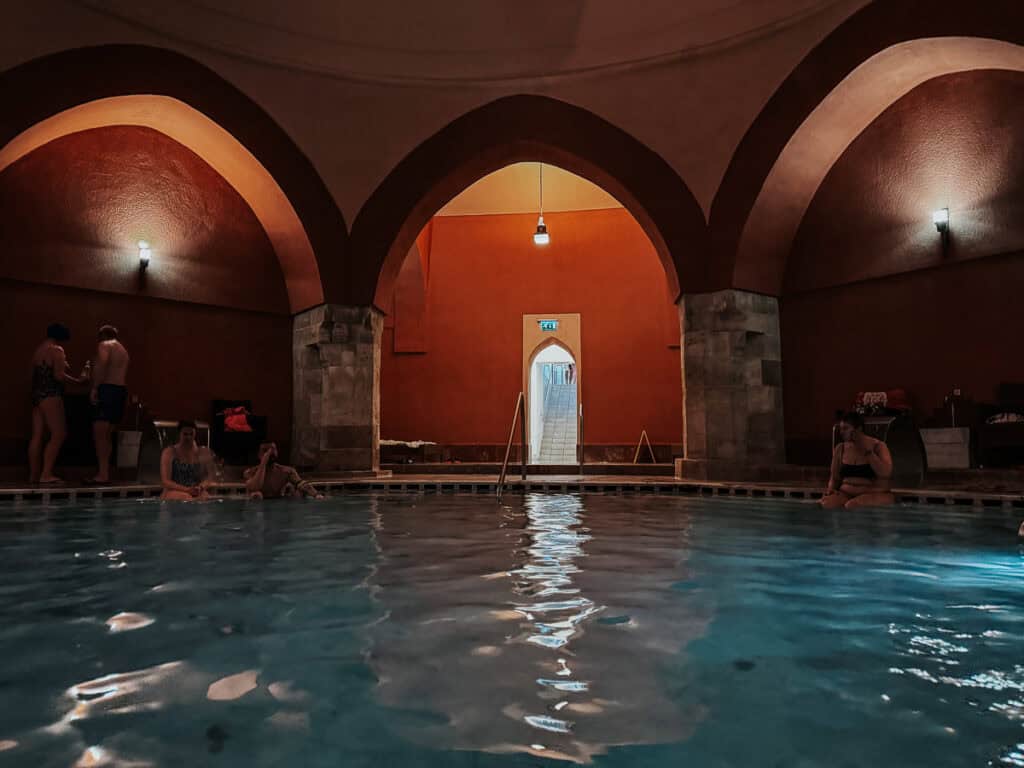 People relaxing and conversing by the indoor pool of Veli Bej Baths, with warm lighting and arched ceilings creating a cozy atmosphere.