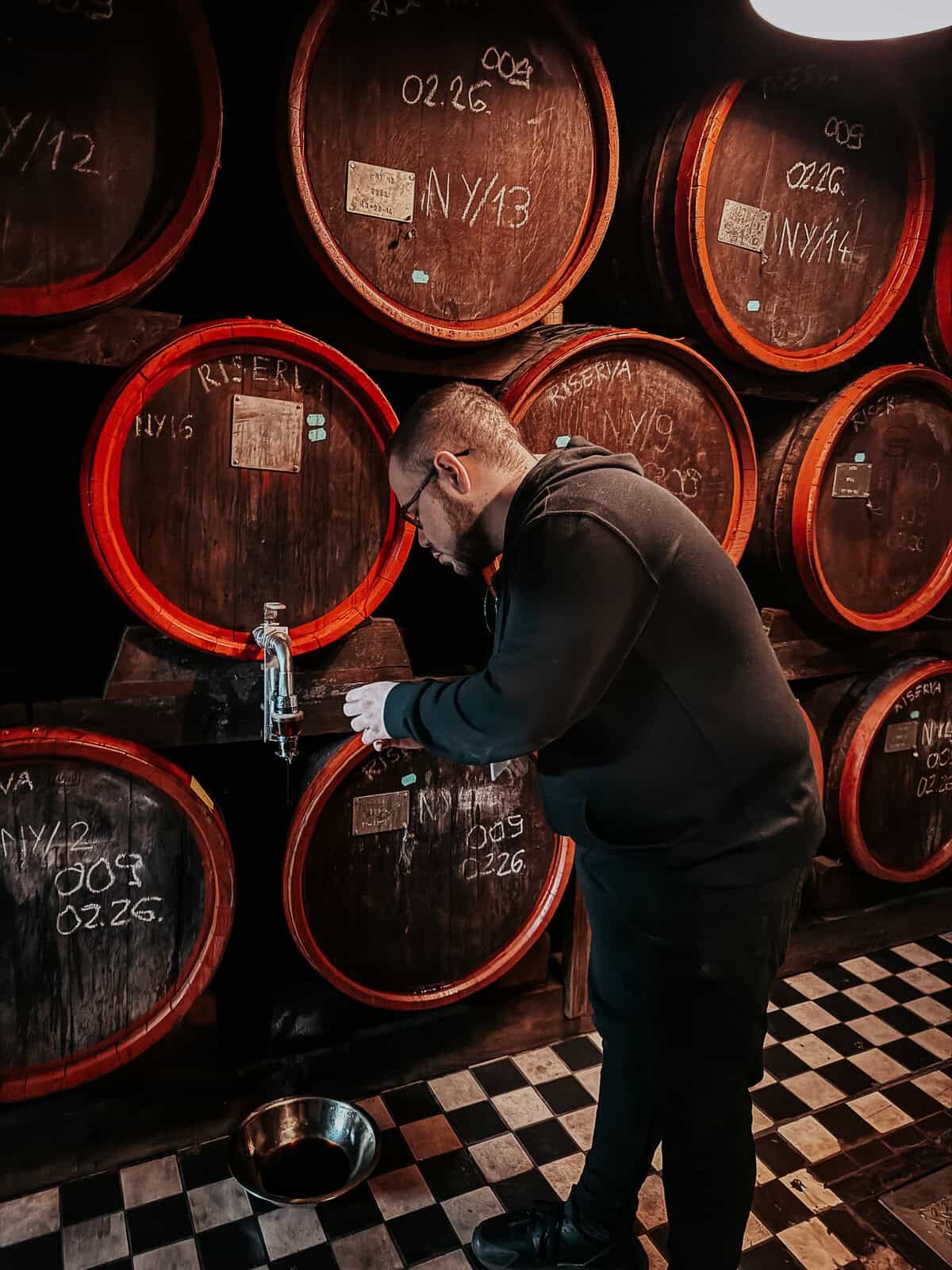 A man in a black hoodie is pouring liquid from a large wooden barrel in the Zwack Museum. The barrels are labeled with various codes and dates, and the floor has a black and white checkered pattern