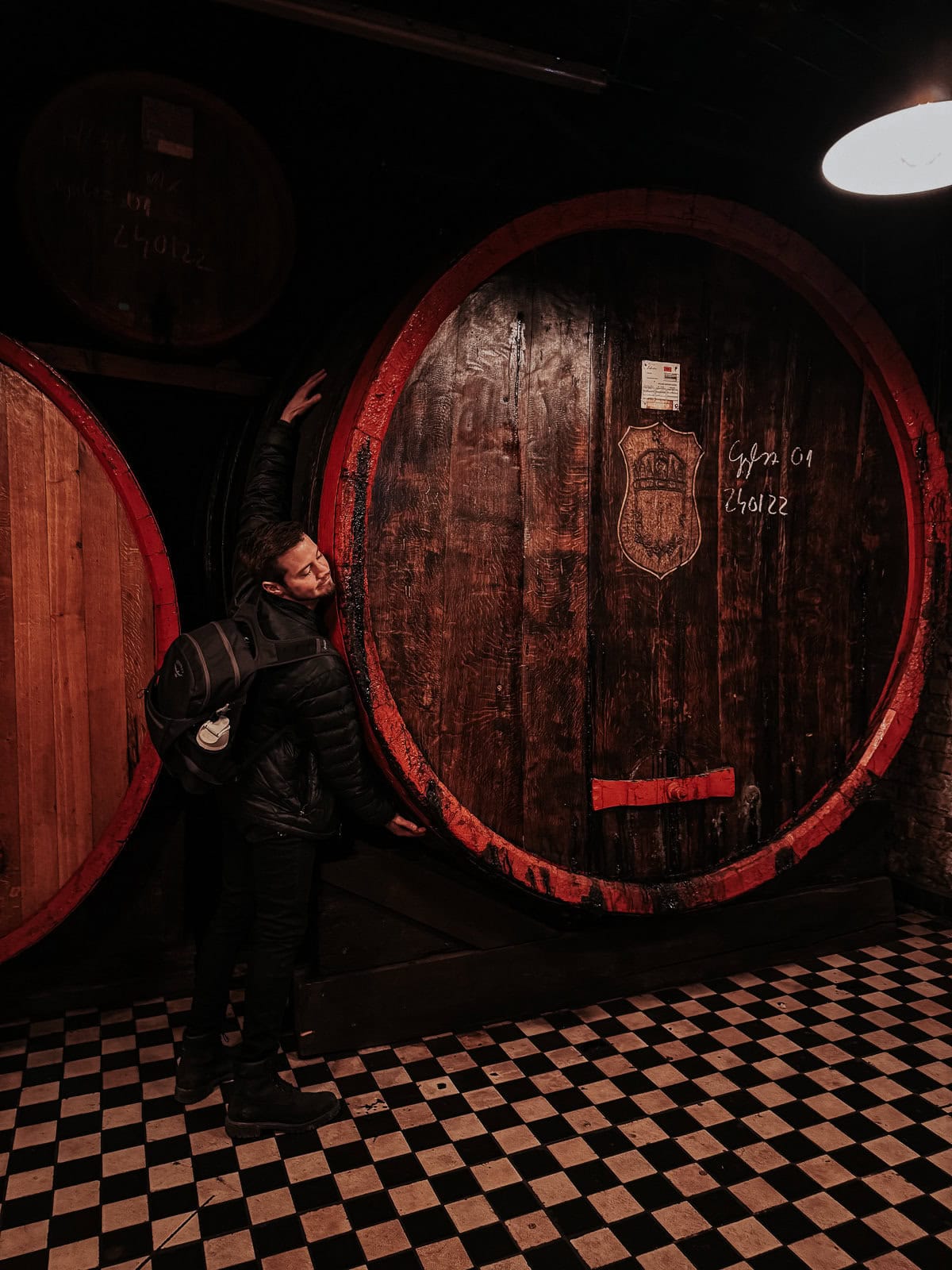 A man with a backpack is hugging a gigantic wooden barrel in the Zwack Museum. The barrel is part of a collection, and the floor is tiled with a black and white checkerboard pattern