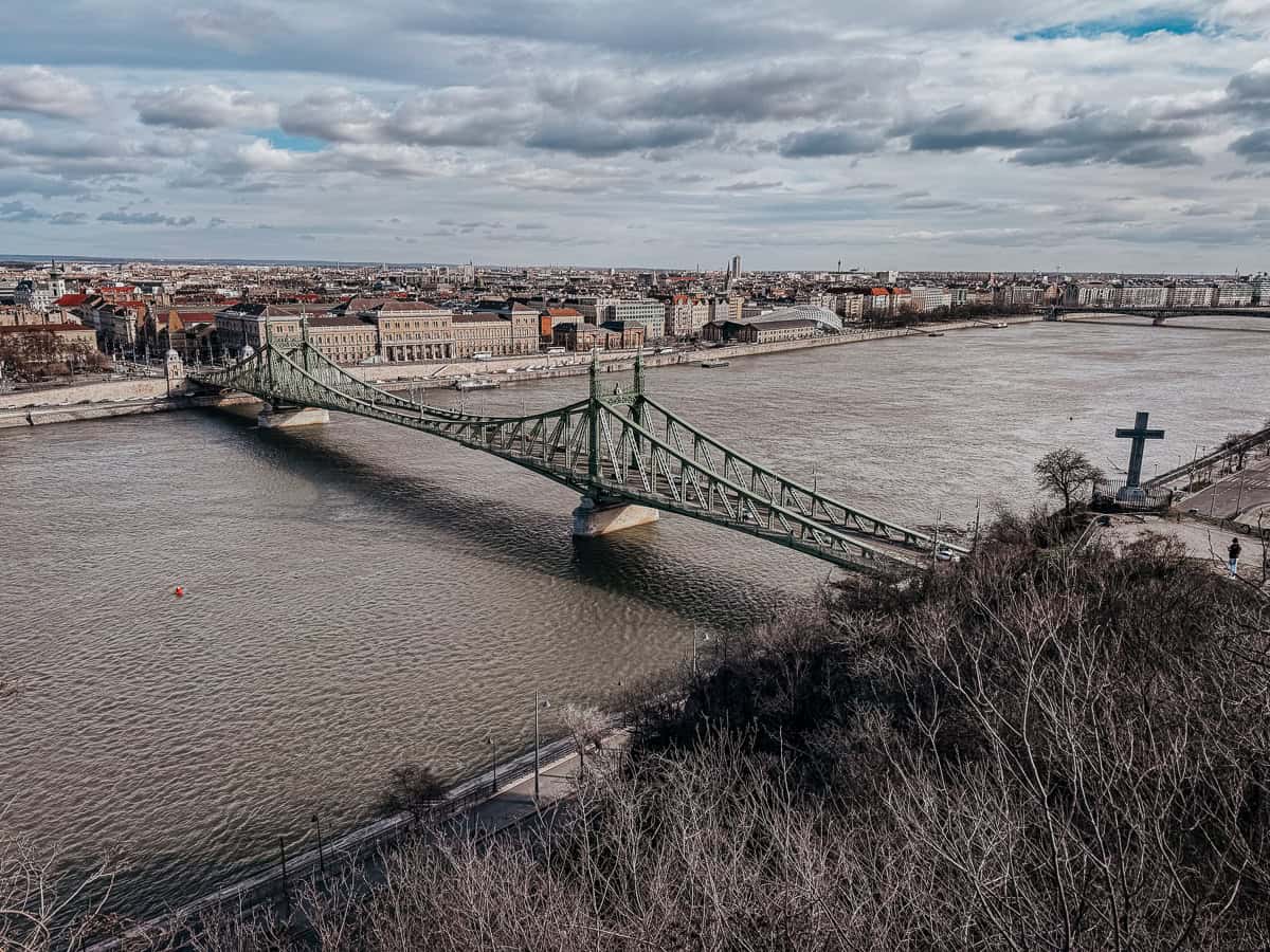 A view of the Liberty Bridge spanning the Danube River, with the cityscape of Budapest in the background under a partly cloudy sky