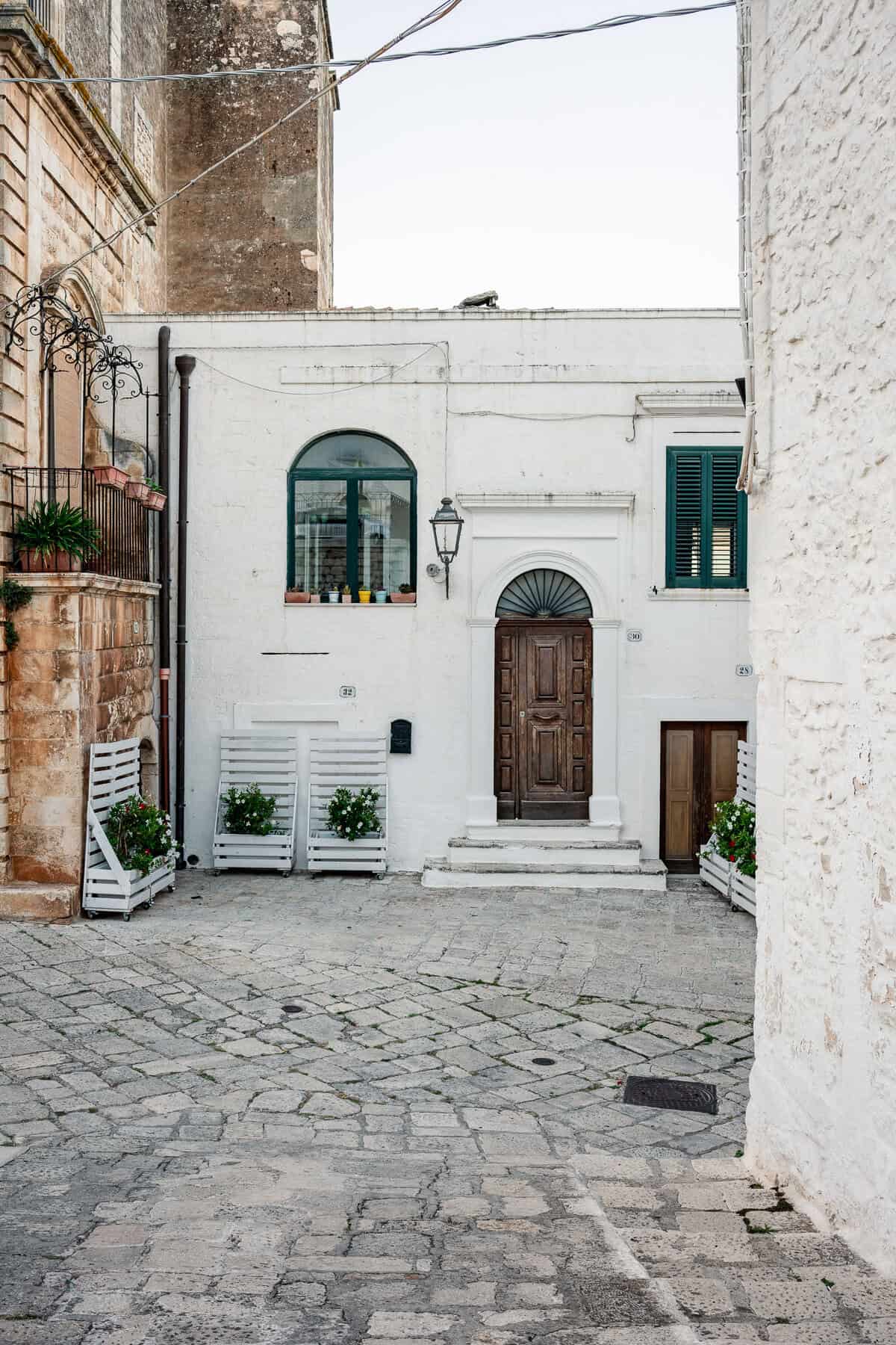 A close-up of a picturesque doorway, decorated with potted flowers, green shutters, and an aged wooden door.